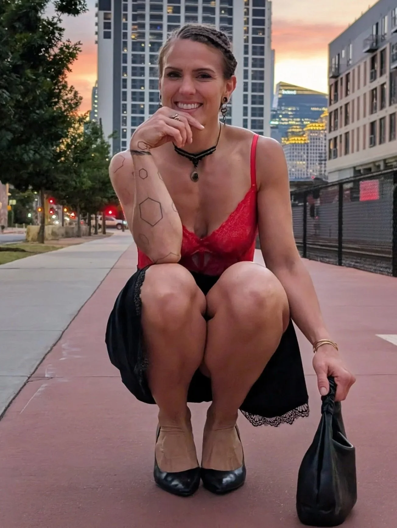 A woman crouching on a city sidewalk during sunset, wearing a red lace top, black skirt, black heels, and holding a black bag.