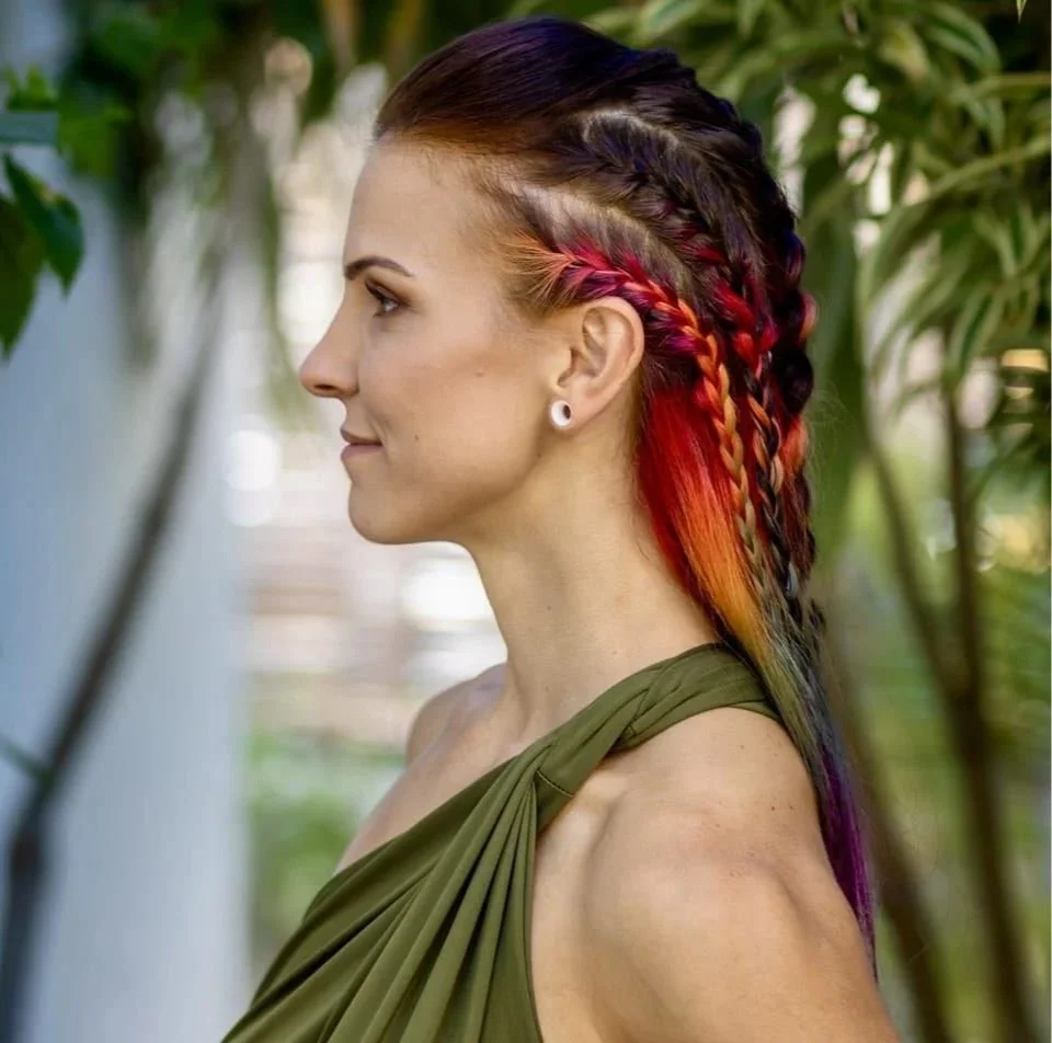 Side profile of a woman with multi-colored braided hair, wearing a green one-shoulder top, standing outdoors with green foliage in the background.