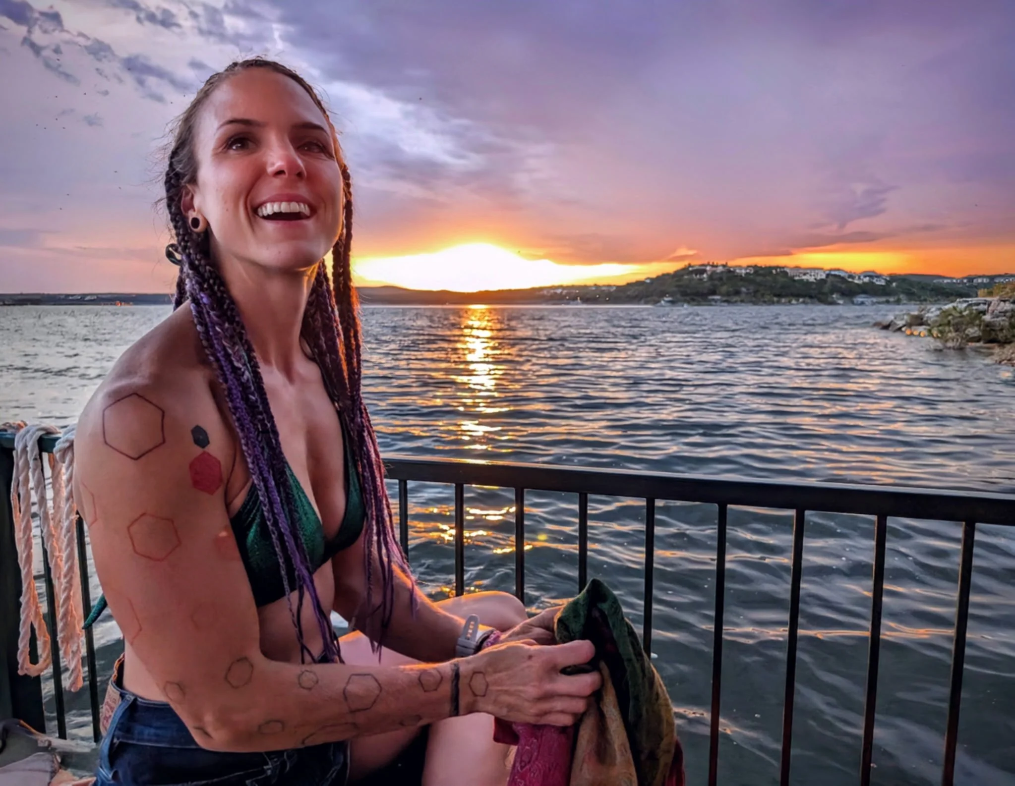 A woman with dreadlocks and tattoos on her arm, smiling while sitting on a boat railing at sunset by the water, with a hill and houses in the background.