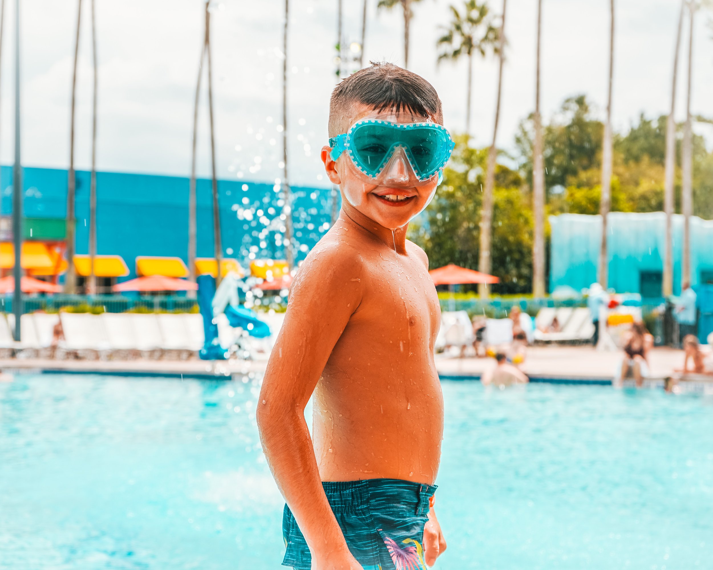 Boy with swim goggles near a pool smiling, with palm trees and people lounging around the pool area.