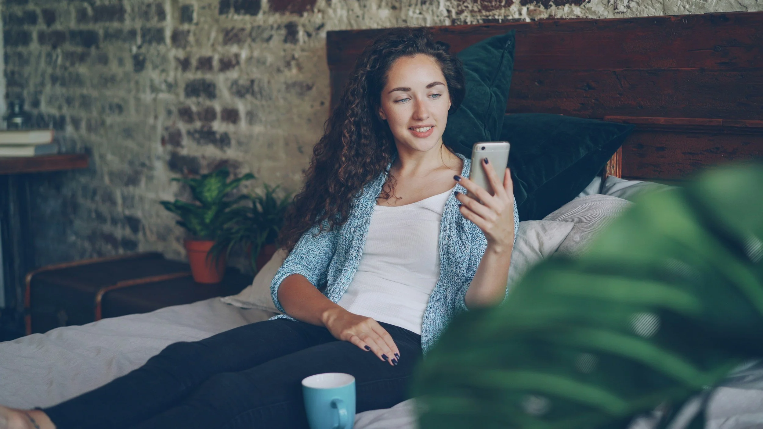 A young woman with curly hair is lying on a bed looking at her smartphone. She has a smile on her face and is dressed casually. There are green plants and a brick wall in the background, and a mug is on the bed next to her.