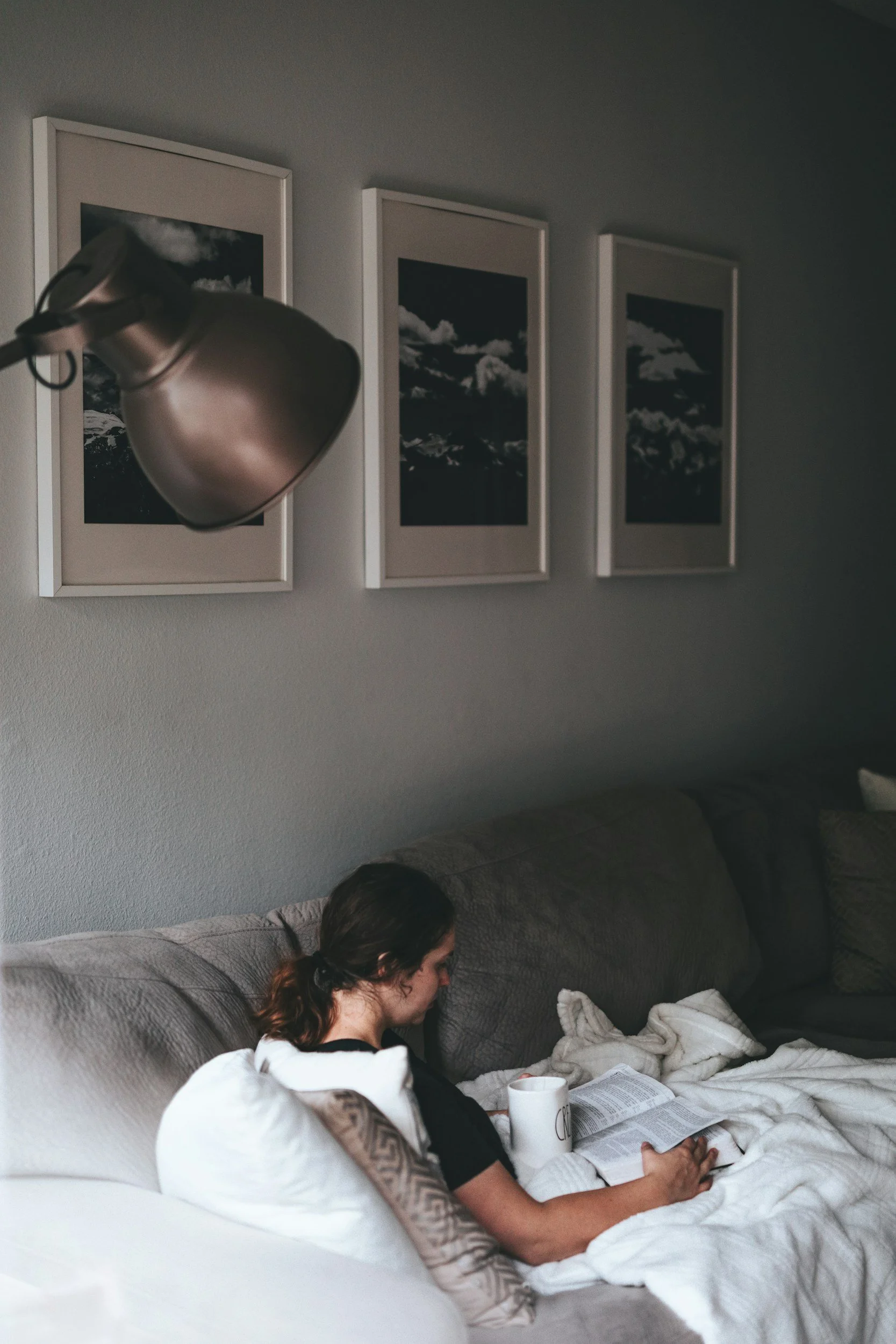 A woman with tattoos on her arm, sitting on a beige couch, reading a newspaper, with a white pillow behind her and a white blanket wrapped around her legs, in a living room with three framed black and white cloud landscapes on the wall above.