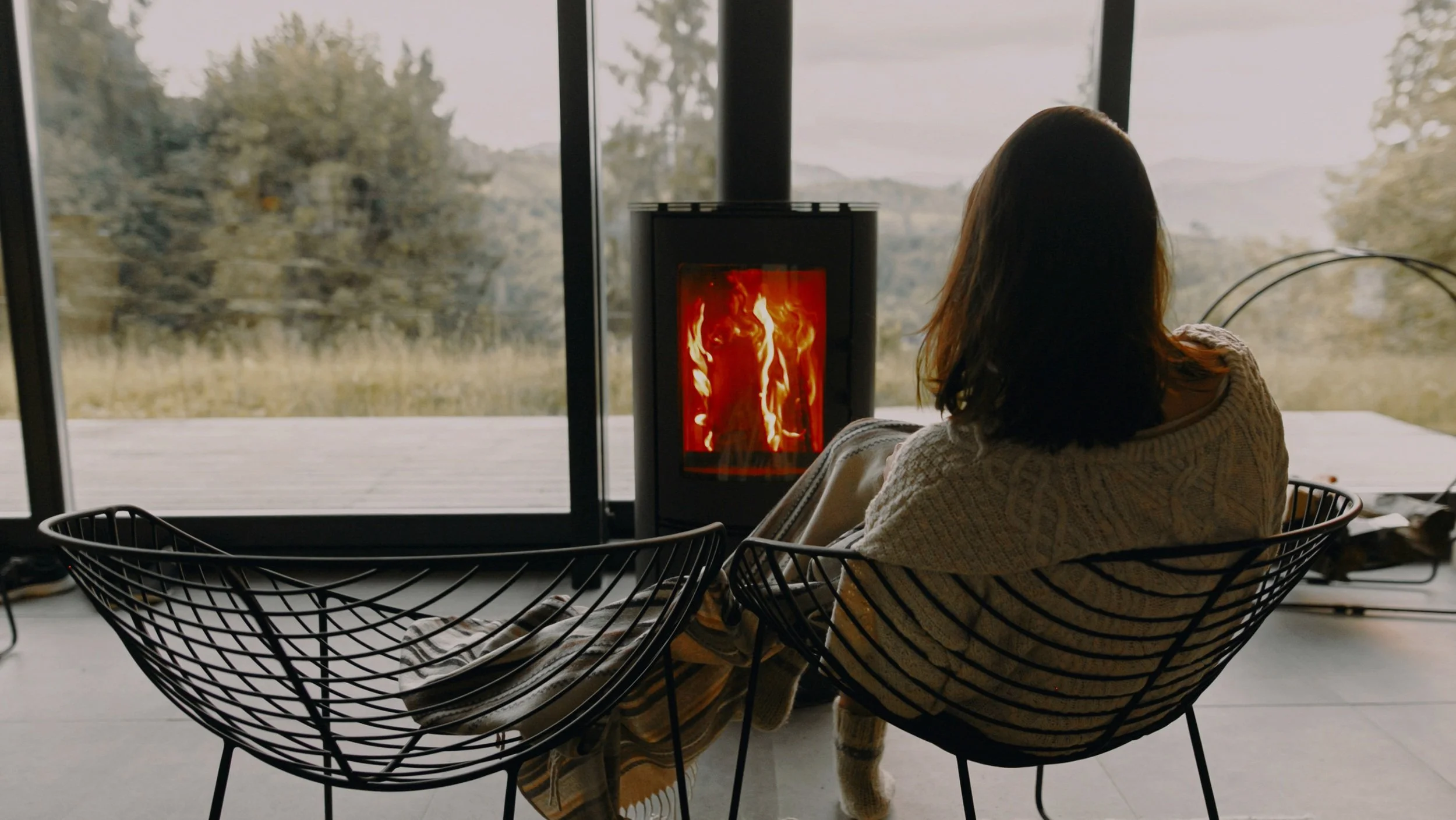 A woman with brown hair sitting in a modern black wire chair in front of large glass windows, looking outside at trees and rolling hills on a cloudy day, with a book on her lap and a small wood stove burning inside.