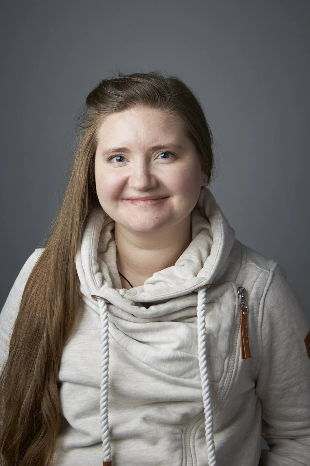 A young woman with long brown hair, smiling, wearing a beige hoodie with a cowl neck and drawstrings, against a gray background. An Online Chronic pain counsellor in canada.