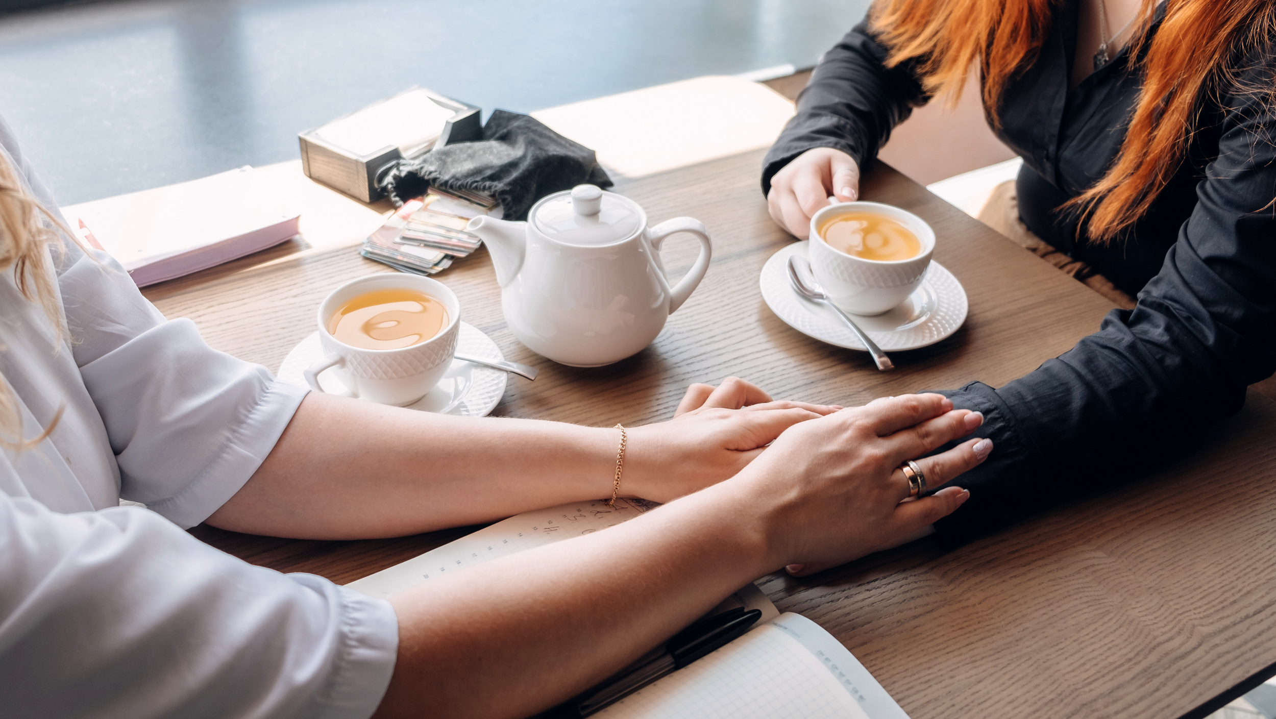 Two women holding hands across a wooden table with tea cups, a teapot, and notebooks. Meeting clients where they're at in their chronic pain journey.
