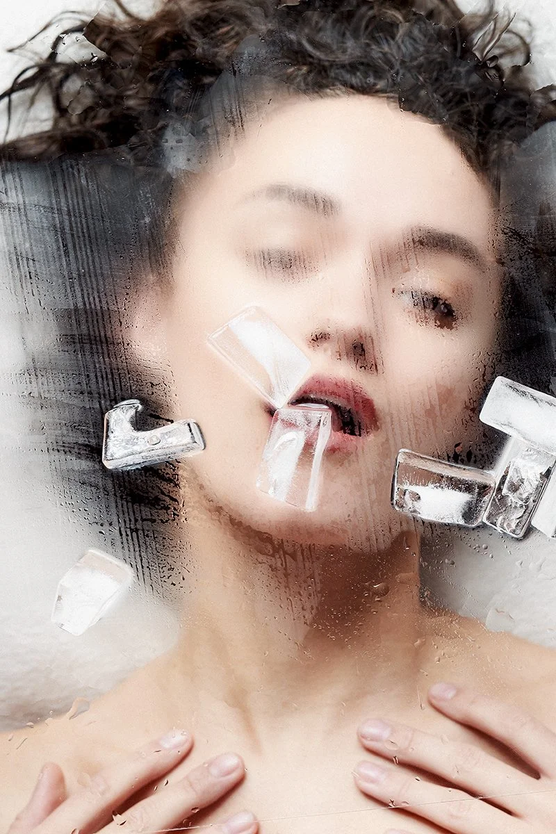 Eine Frau mit nasser Haut und geschlossenen Augen, hinter einer Glas- oder Plastikscheibe mit Wassertröpfchen, bedeckt mit Eiswürfeln.