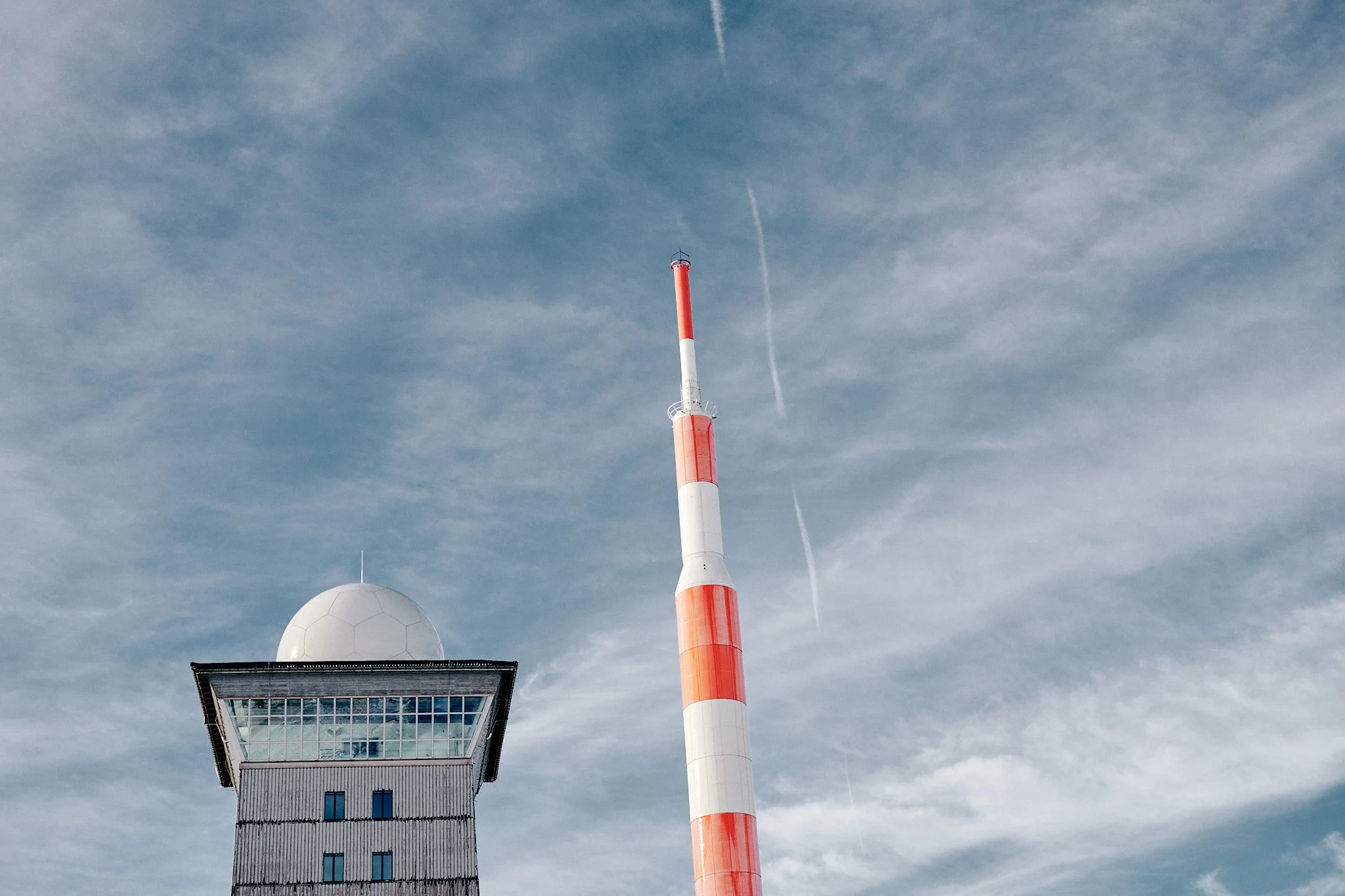 Ein Fernmeldeturm mit roter und weißer Bemalung und einem Windrad im unteren Bereich, vor einem bewölkten Himmel mit Kondensstreifen.