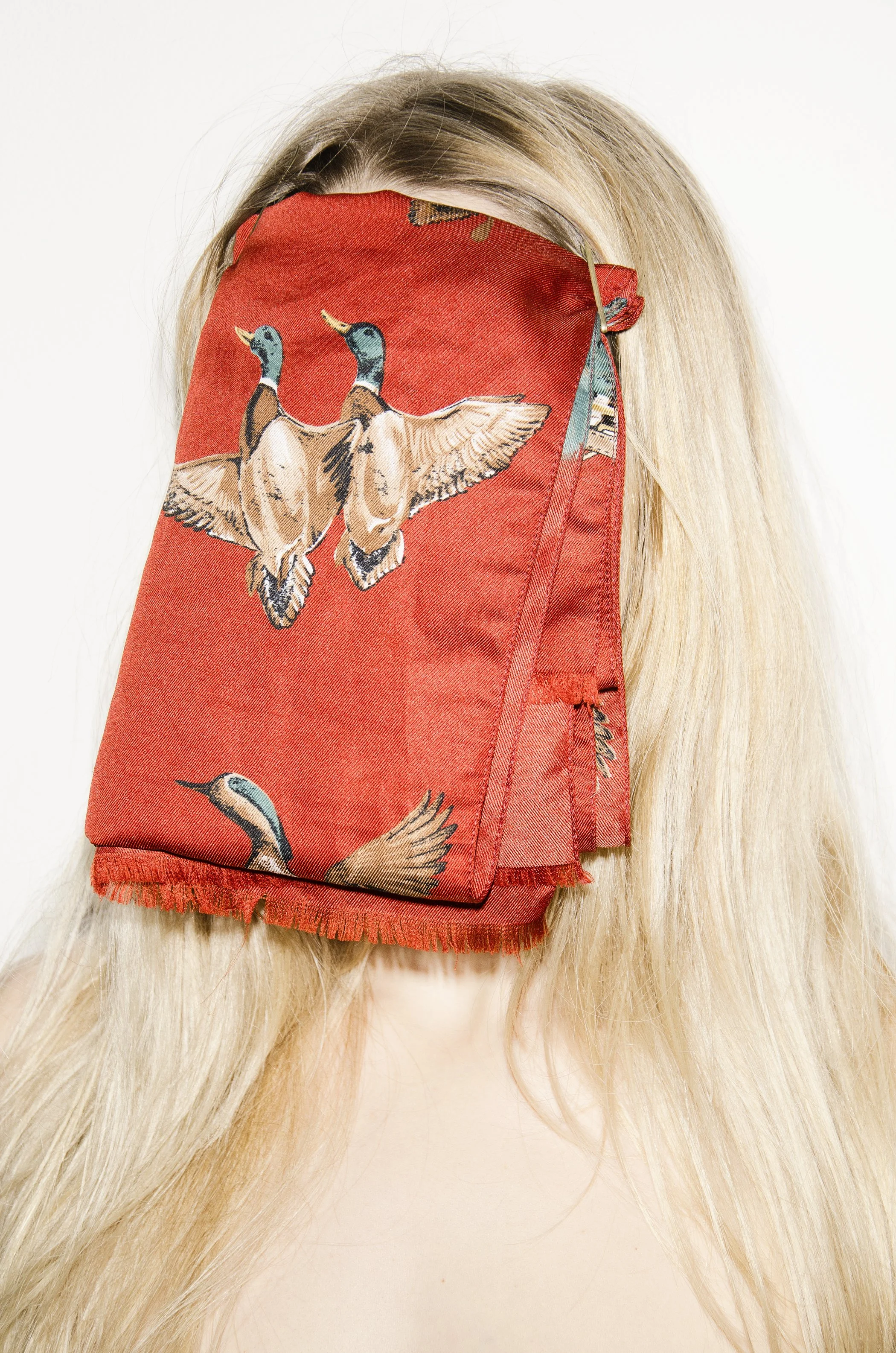 Eine Frau mit langem blondem Haar trägt eine rote Stoffmaske mit Entenmotiven vor einem weißen Hintergrund.