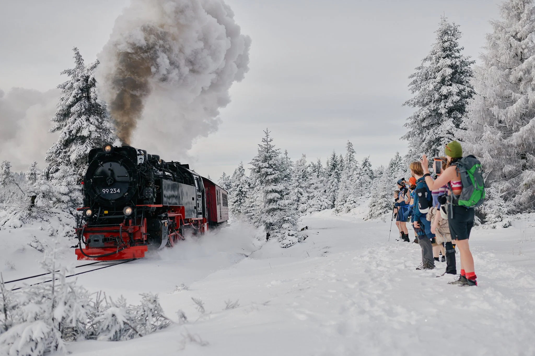 Eine Lokomotive fährt durch eine verschneite Winterlandschaft, während eine Gruppe von leichtbekleideten Erwachsenen am Bahnsteig Fotos macht.