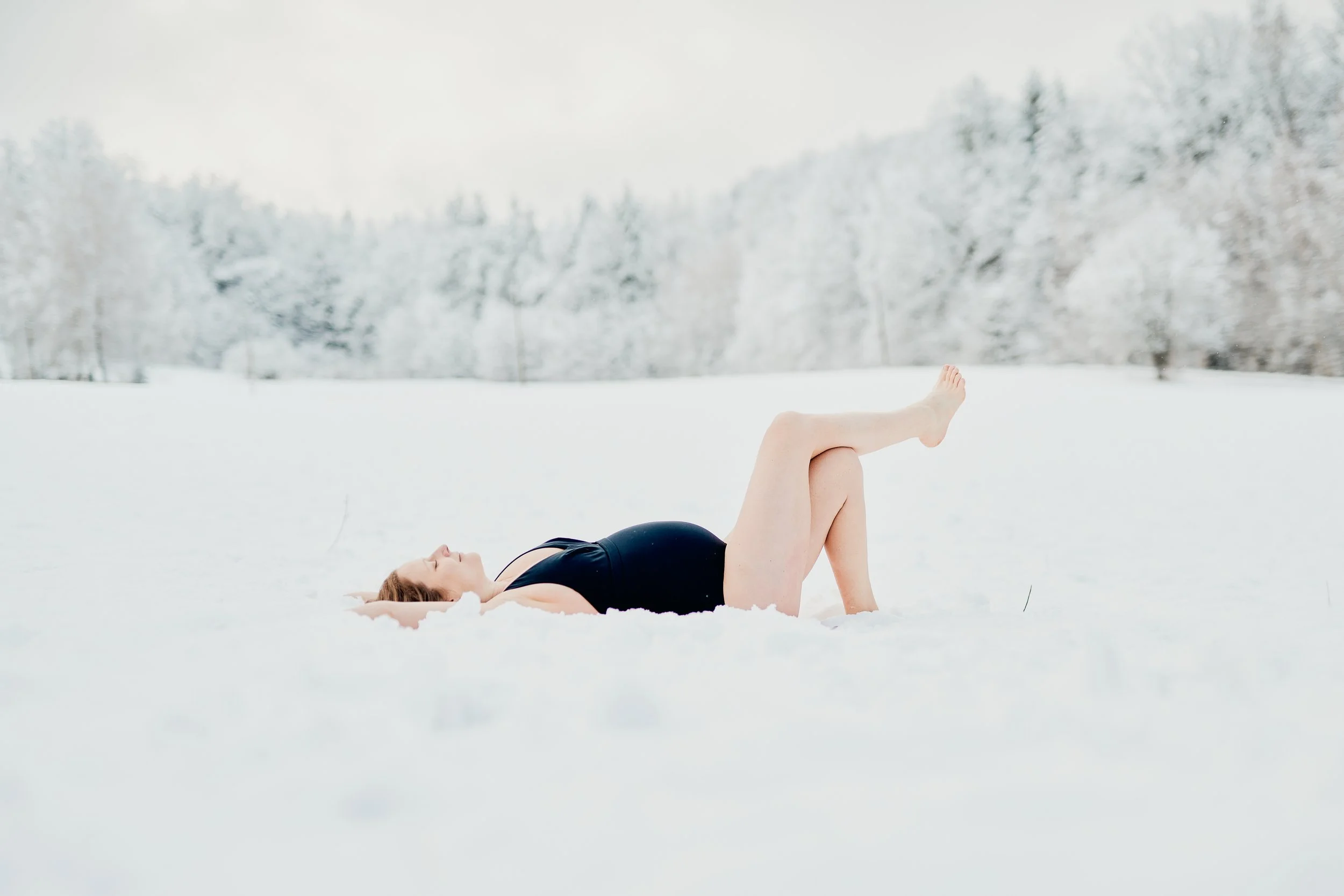 Schwangere Frau liegt entspannt im Schnee, im Hintergrund verschneite Bäume.