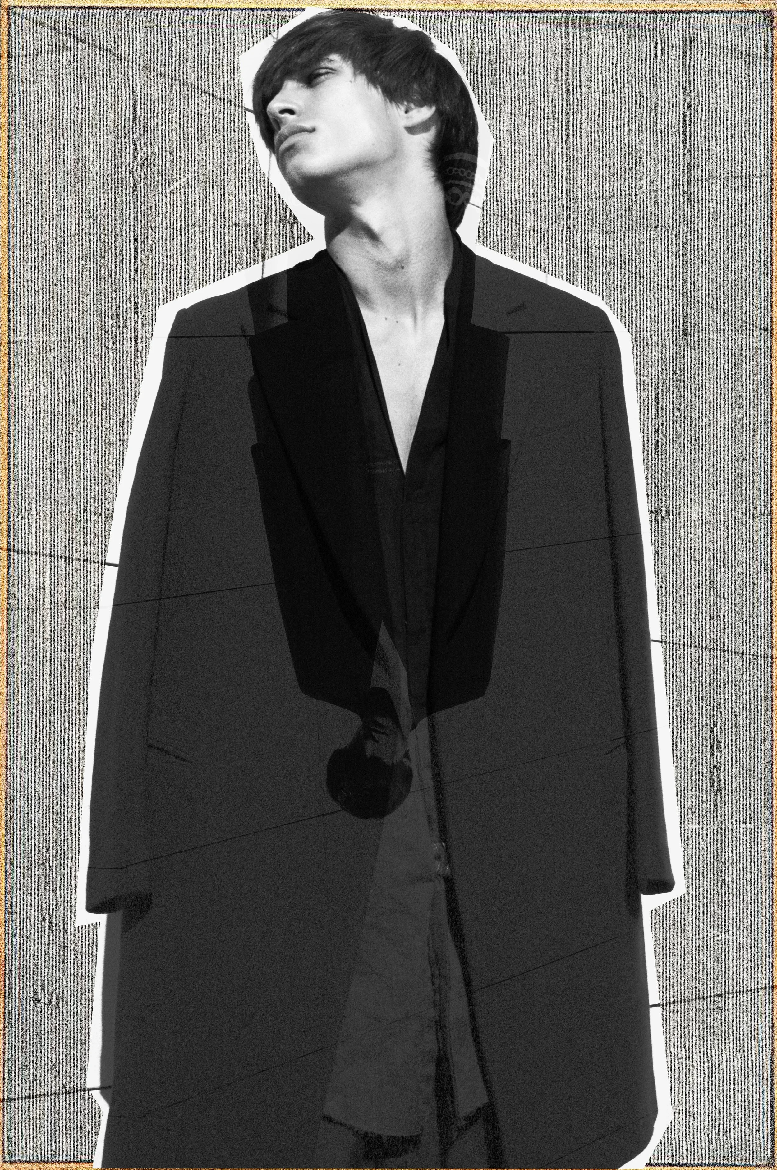 Schwarz-Weiß-Foto einer Person mit kurzem schwarzen Haar, die eine schwarze Jacke und ein schwarzes Hemd trägt, auf einem Hintergrund mit vertikalen Linien.