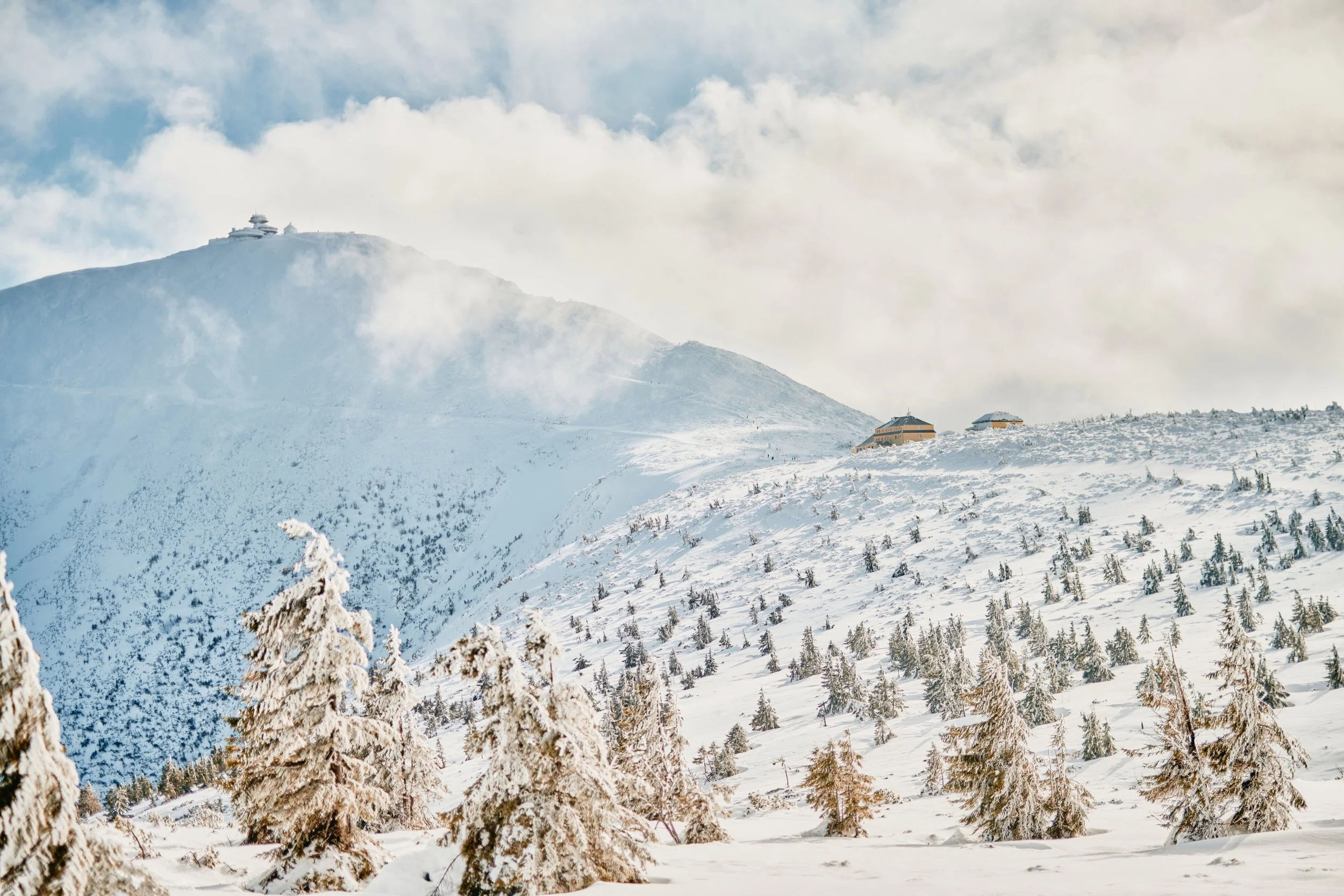Schneebedeuerte Berge und Bäume, einige Gebäude auf dem Berggipfel, bewölkter Himmel.