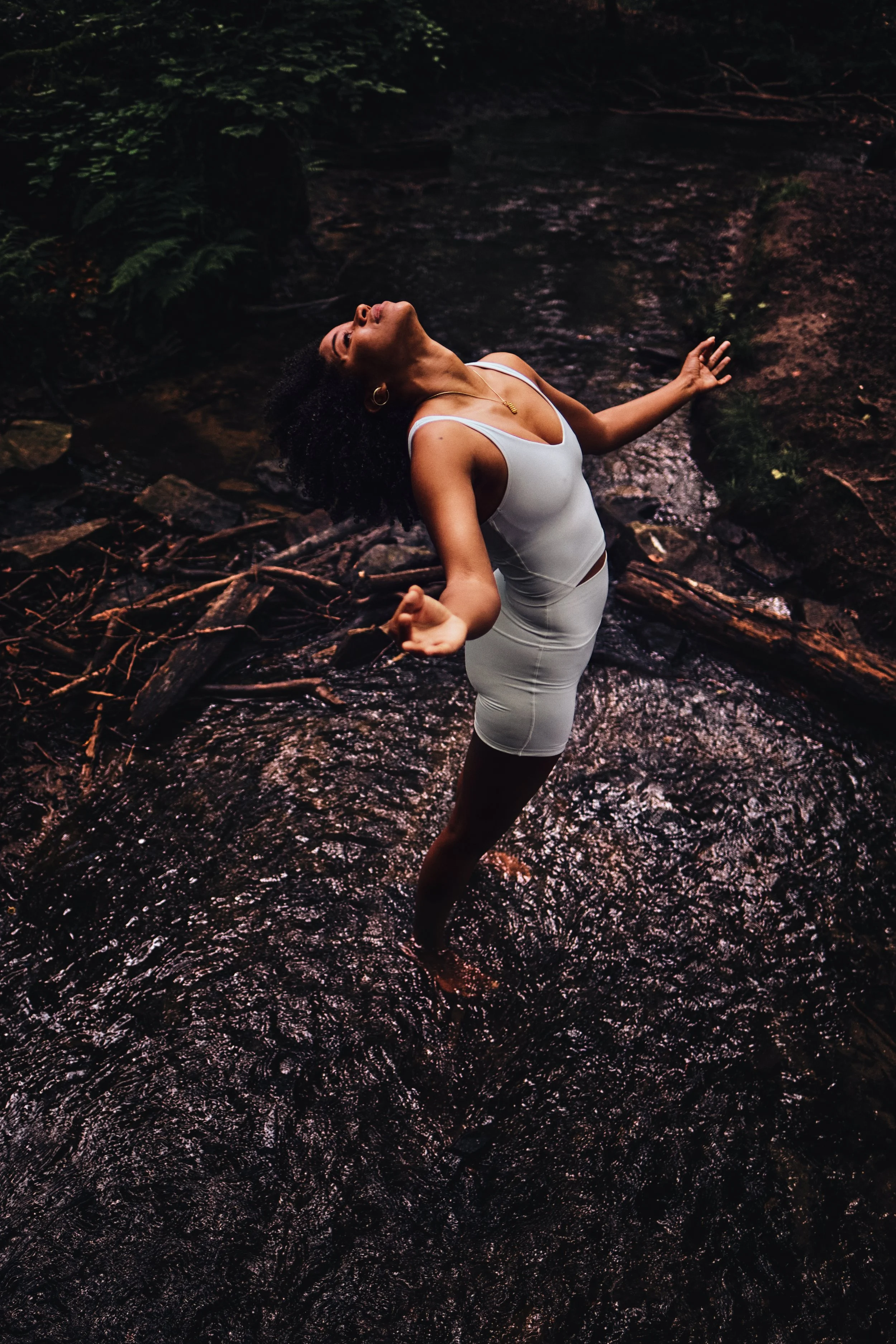 Eine Frau trägt ein weißes Kleid, steht mit geneigtem Kopf und offenem Mund in einem kleinen Fluss inmitten eines Waldes, umgeben von grünen Pflanzen und Baumstämmen.