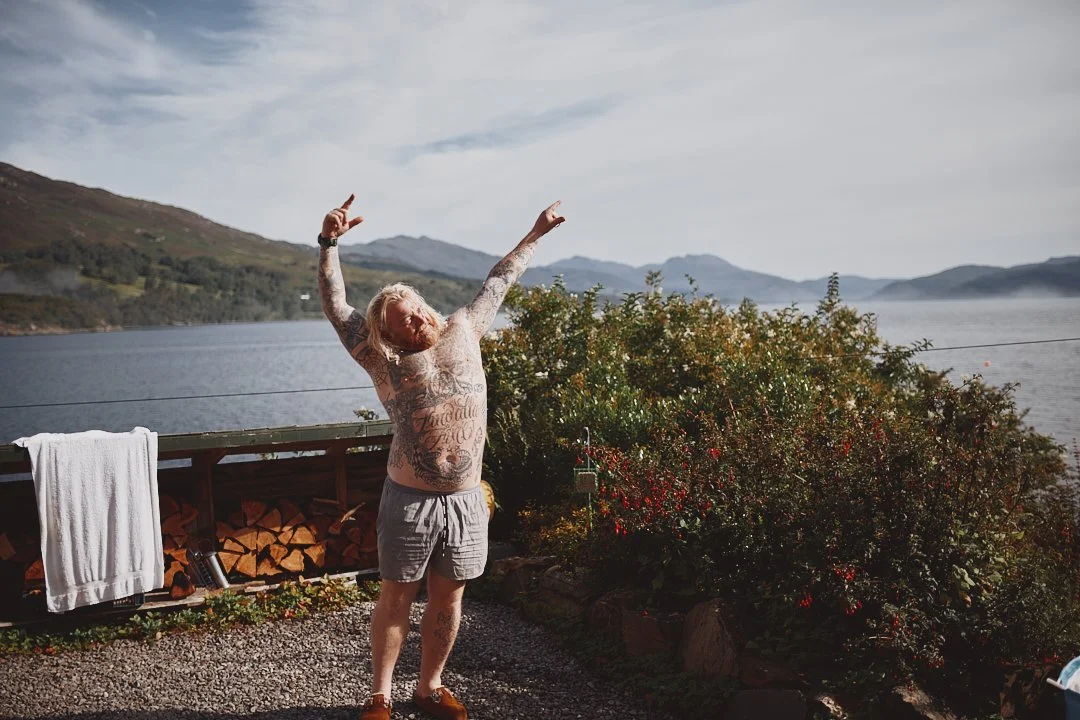 Ein tattooüberzogener Mann mit kurzen Haaren steht vor einem See, hebt die Arme in die Luft und genießt die Natur. Im Hintergrund sind Berge und Wolken am Himmel zu sehen.