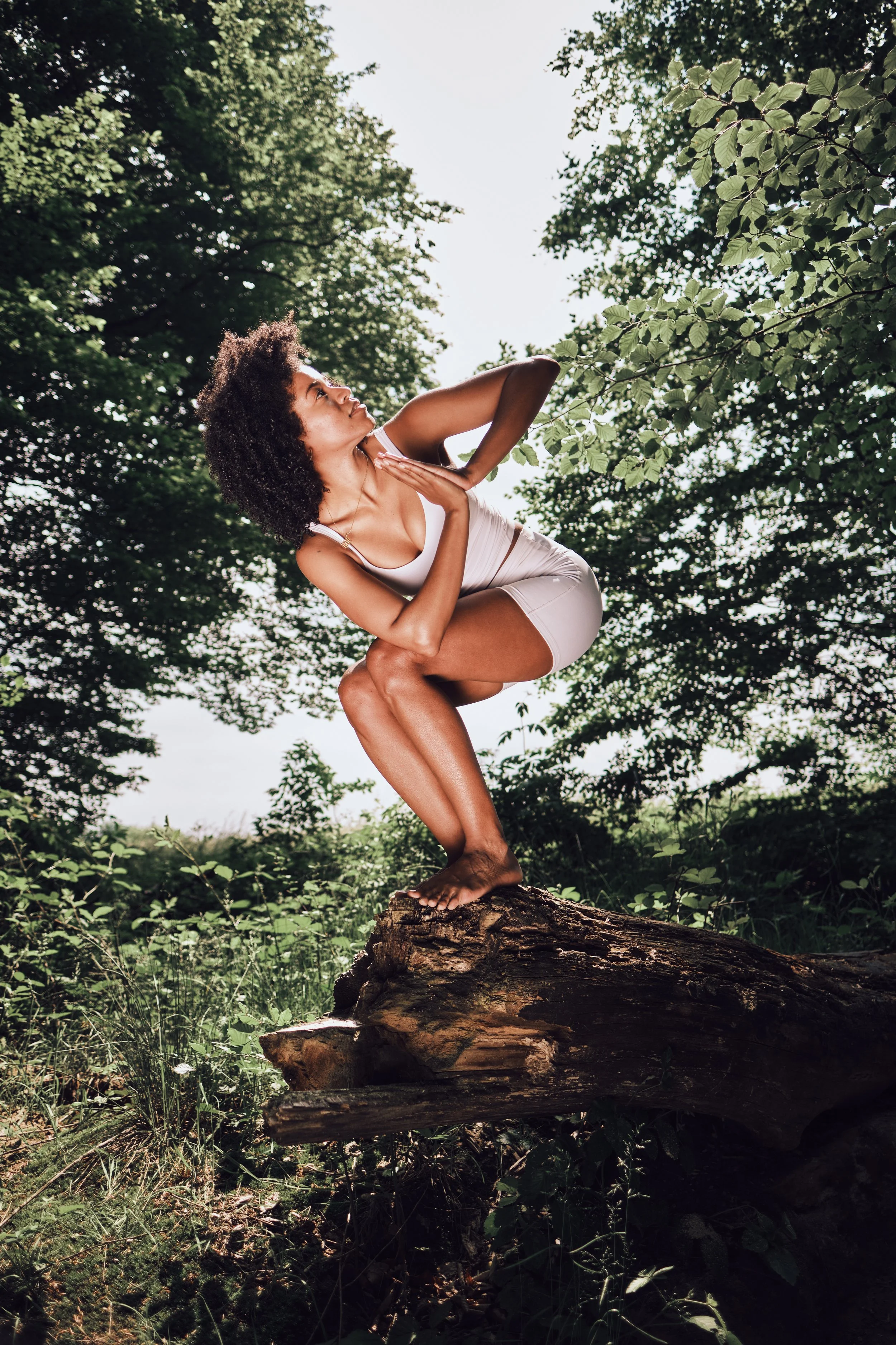 Eine junge Frau in weißem Outfit macht Yoga auf einem Baumstamm im Wald, umgeben von Bäumen und Grünflächen.