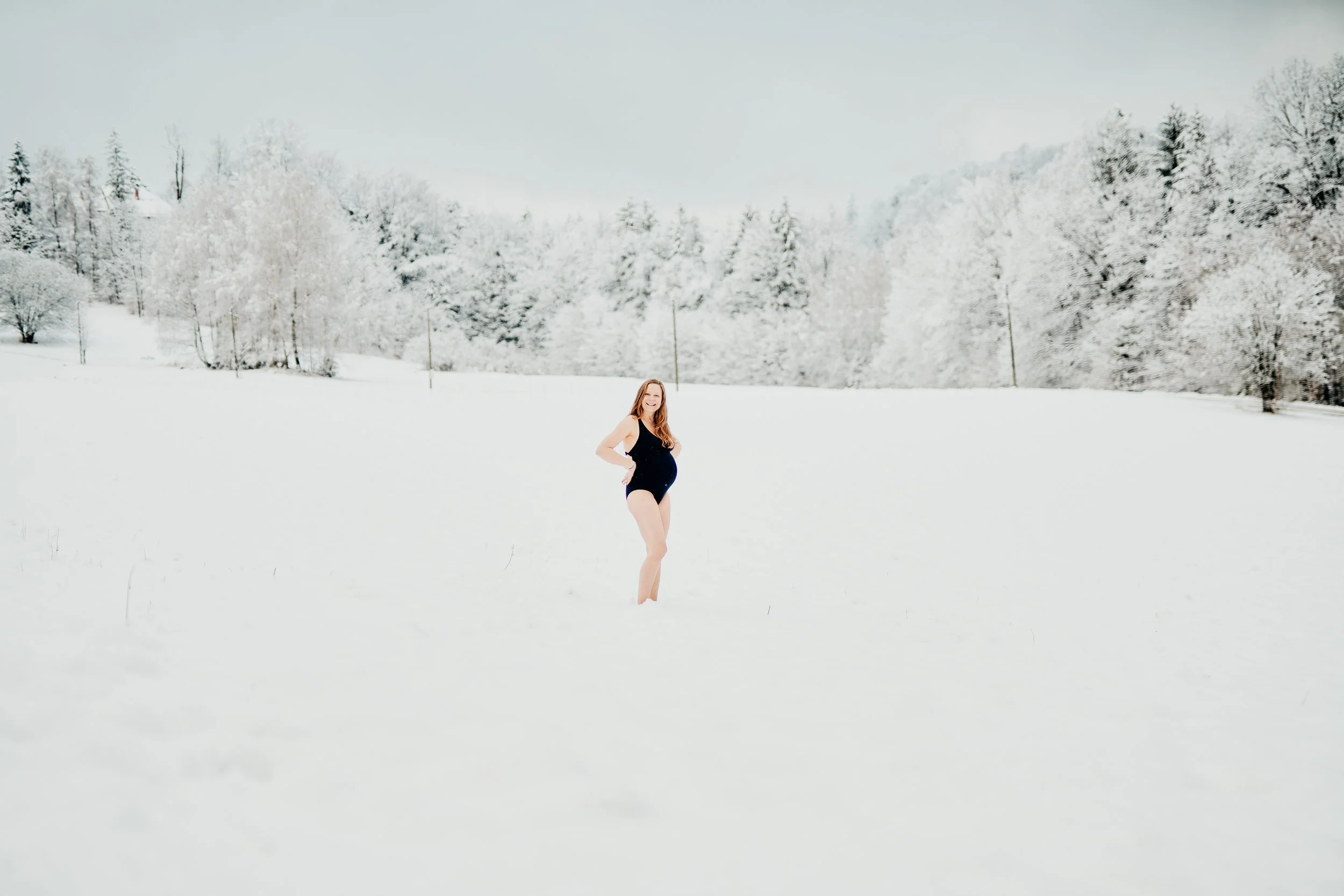 Schwangere Frau im Schnee in einem schwarzen Badeanzug, im Hintergrund verschneite Bäume und ein bewölkter Himmel.