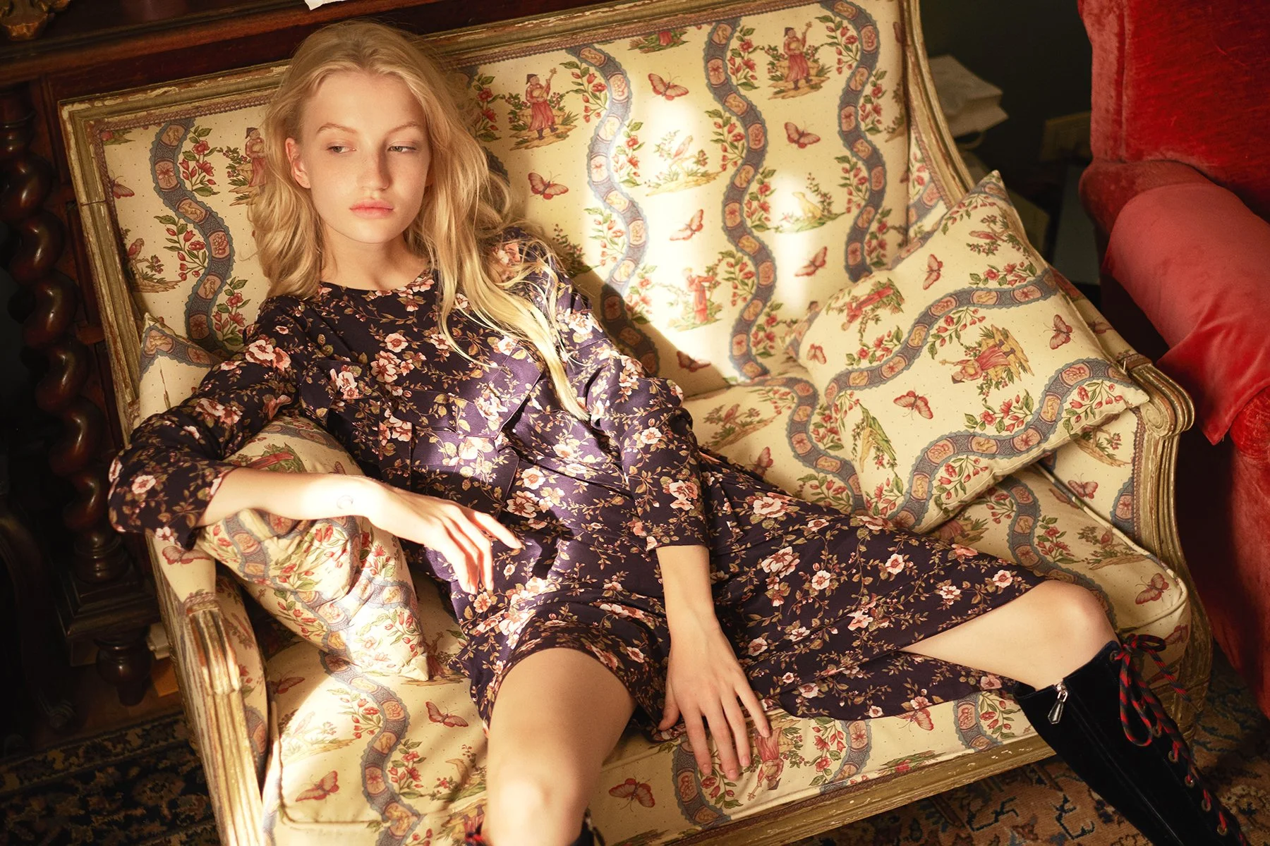 Junge Frau mit blonden Haaren liegt auf einem antiken, gemusterten Sofa, trägt ein dunkles, florales Kleid, eine schwarze Stiefel. Die Einrichtung ist nostalgisch mit geschnitztem Holz und bunten Mustern.