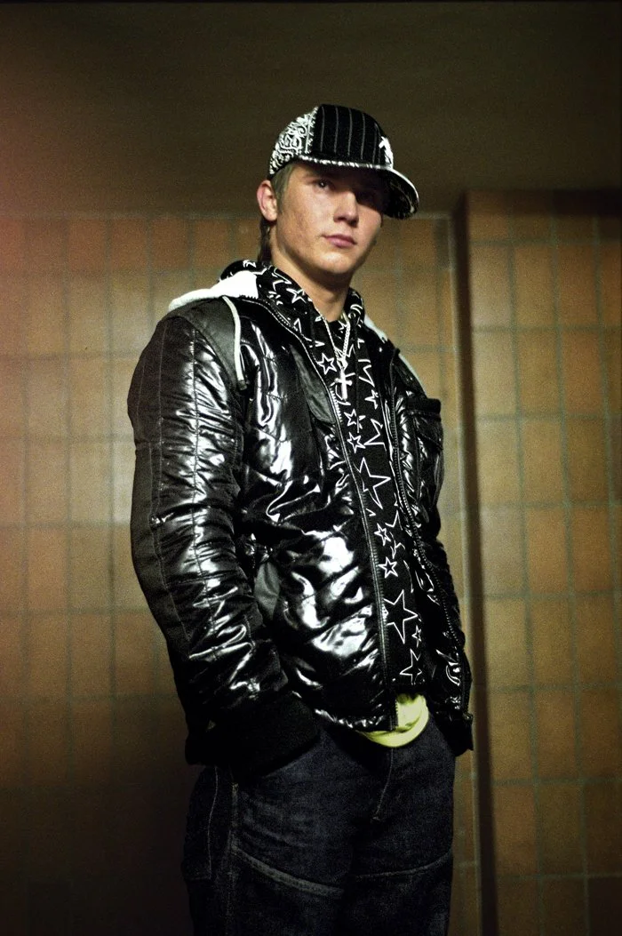 Junge trägt schwarze, glänzende Jacke, Star-Muster-Hoodie, schwarze Kappe mit weißen Mustern, dunkle Hose, in einem Raum mit gestrichenen Wänden.