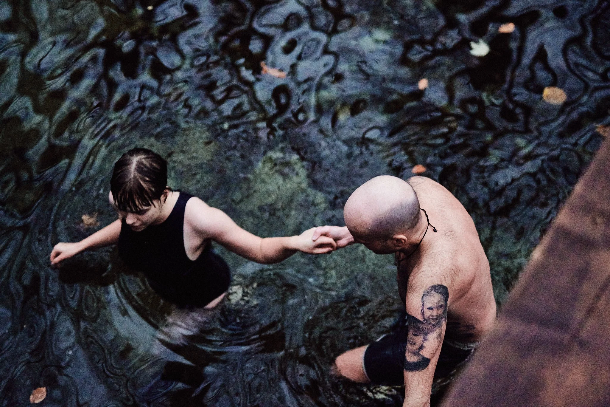 Zwei Menschen stehen in flachem, dunklem Wasser und halten sich an den Händen. Einer hat Tattoos am Arm, der andere trägt eine schwarze Kleidung, und sie schauen sich an.