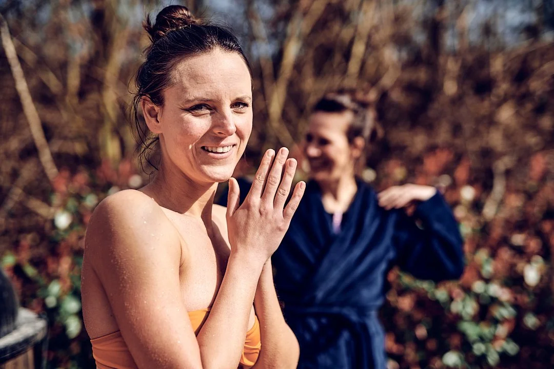 Zwei Frauen im Freien nach einem Wasserbad, die eine lächelnd mit nassen Haaren und die andere mit Hand an der Schulter, im Hintergrund Bäume mit herbstlichen Blättern.