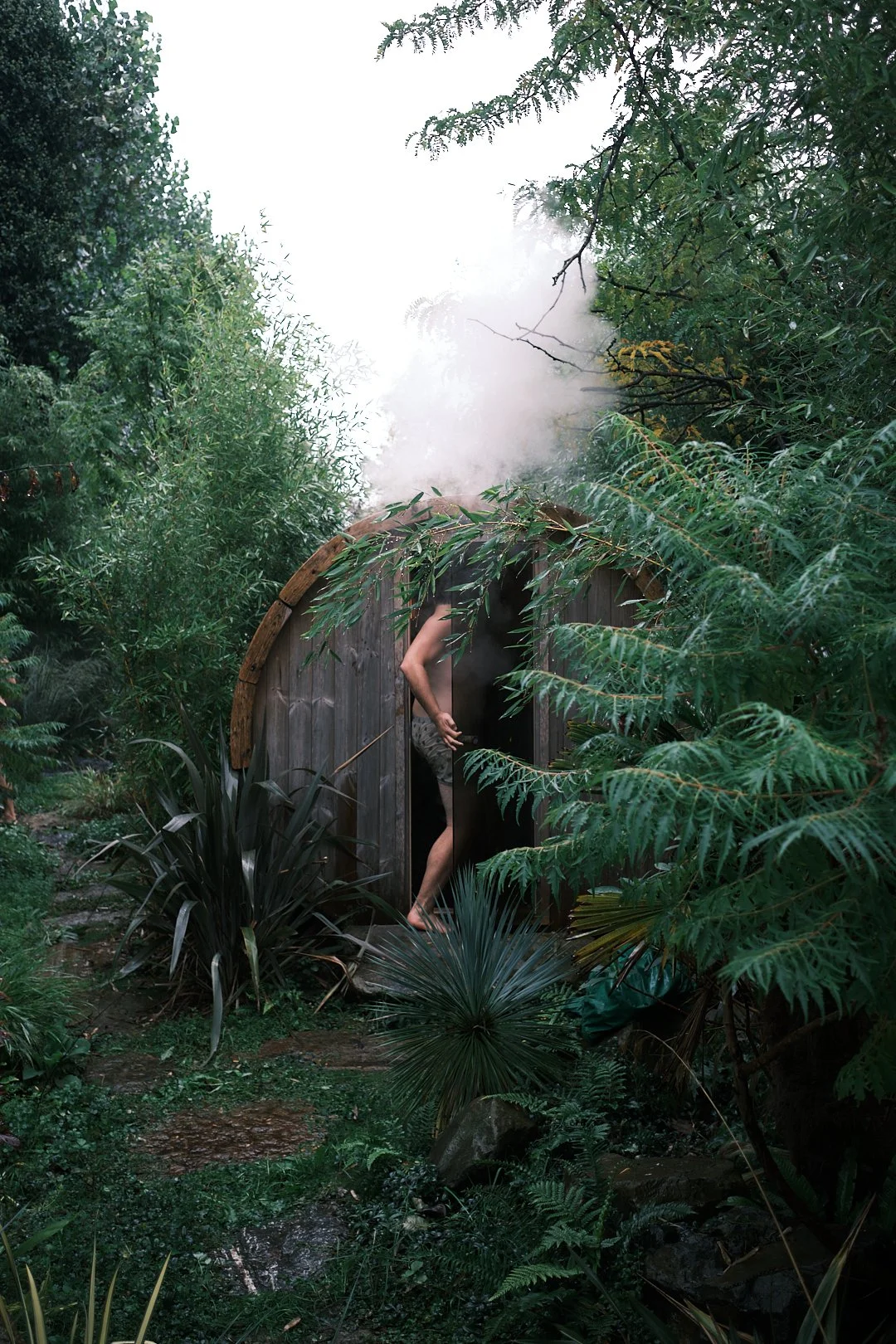 Junge Person geht in eine Fasssaune mit Holz, umgeben von grünen Pflanzen und Bäumen im Wald, mit Nebel oder Dampf im Hintergrund.