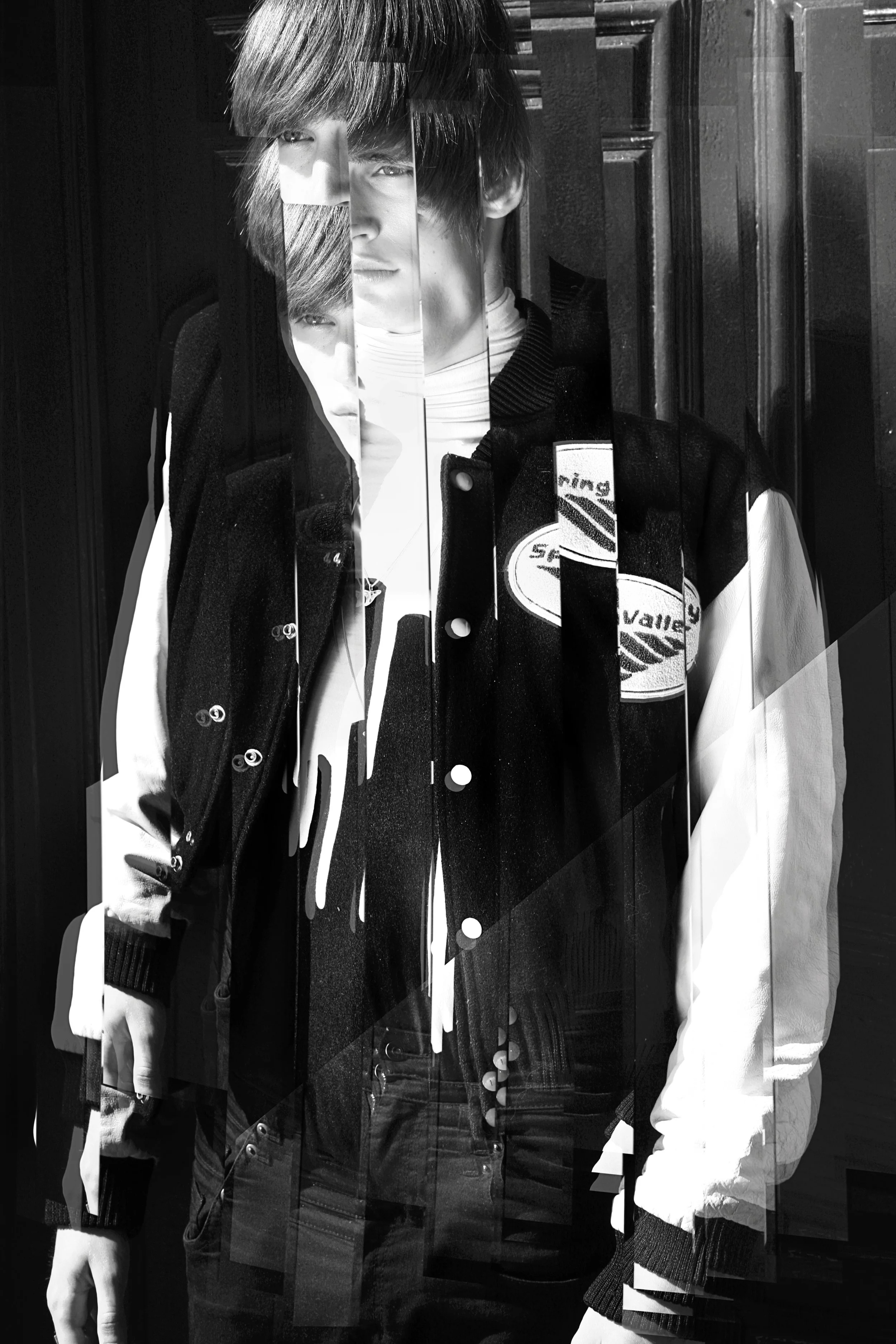 Ein durch Digitalefehler gestörter schwarz-weißer Porträtfoto eines jungen Mannes in einer Jacke mit Aufnähern, der vor einer dunklen Holztür steht.