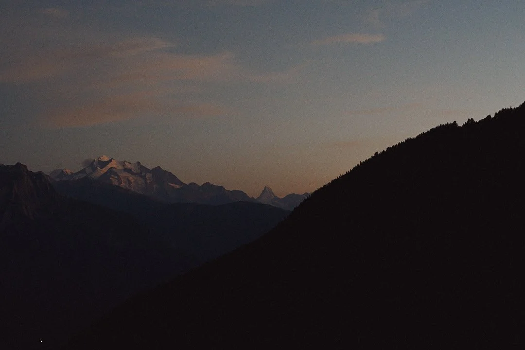 Bergpanorama bei Sonnenuntergang mit schneebedeckten Gipfeln und dunklen Hügeln im Vordergrund.