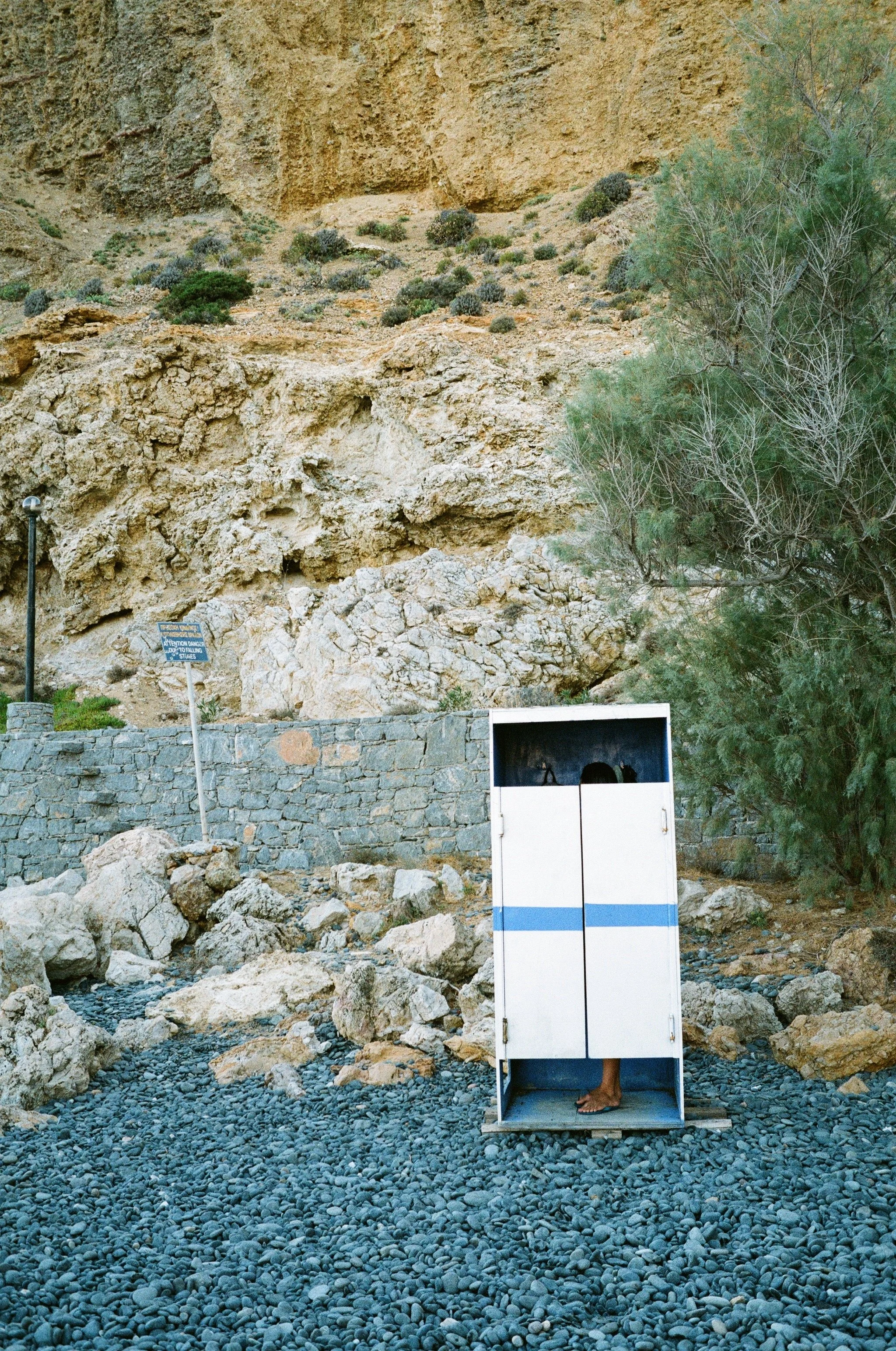 Ein grauer und blauer Kasten mit einem offenen unteren Teil, in dem Füße eines Menschen sichtbar sind, steht auf einem felsigen Boden vor einer felsigen Wand, umgeben von Steinen und einem Baum auf der rechten Seite.