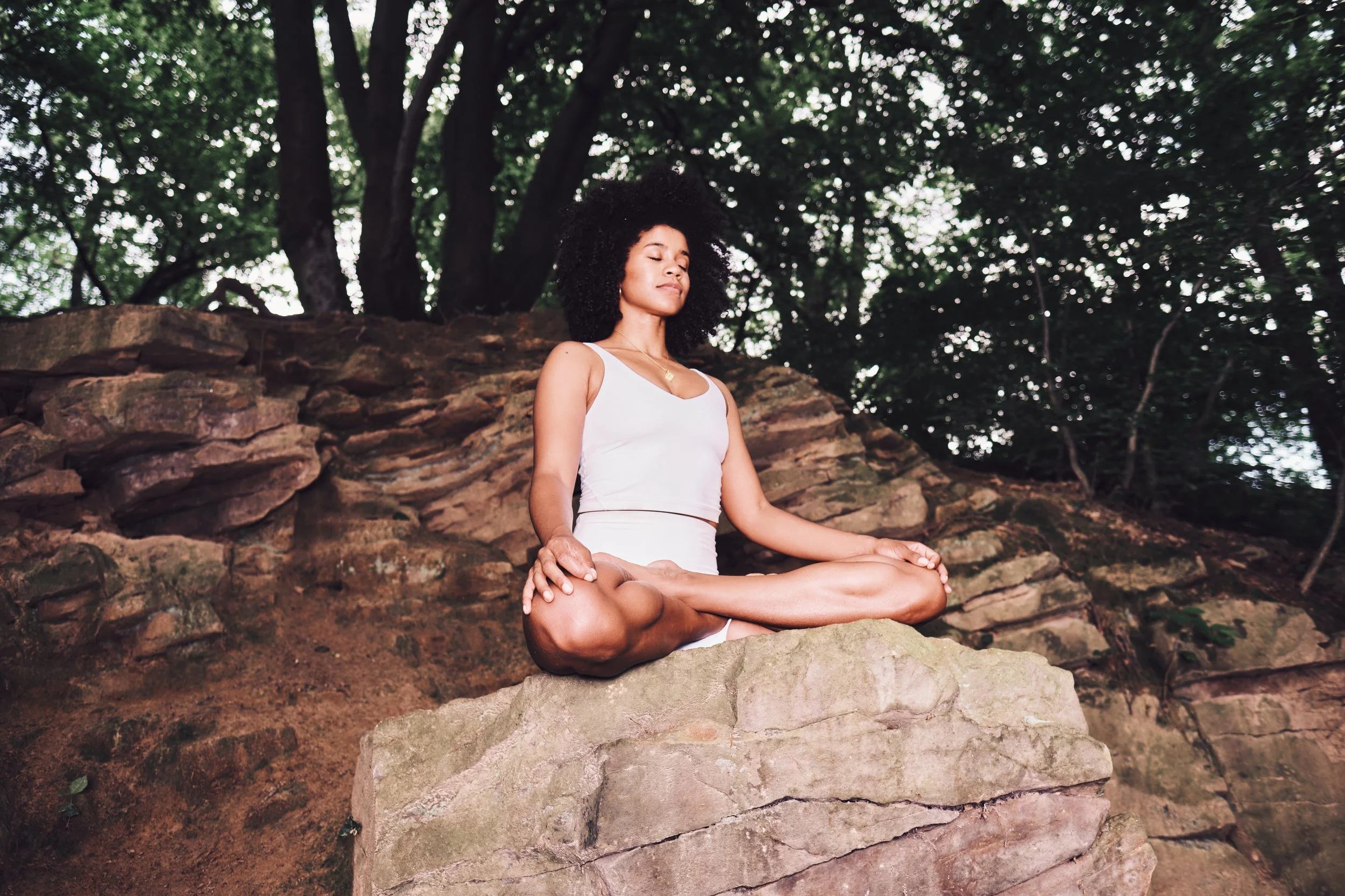 Eine junge Frau sitzt im Schneidersitz auf einem Stein, umgeben von Bäumen, mit geschlossenen Augen, die Meditation oder Entspannung symbolisieren.