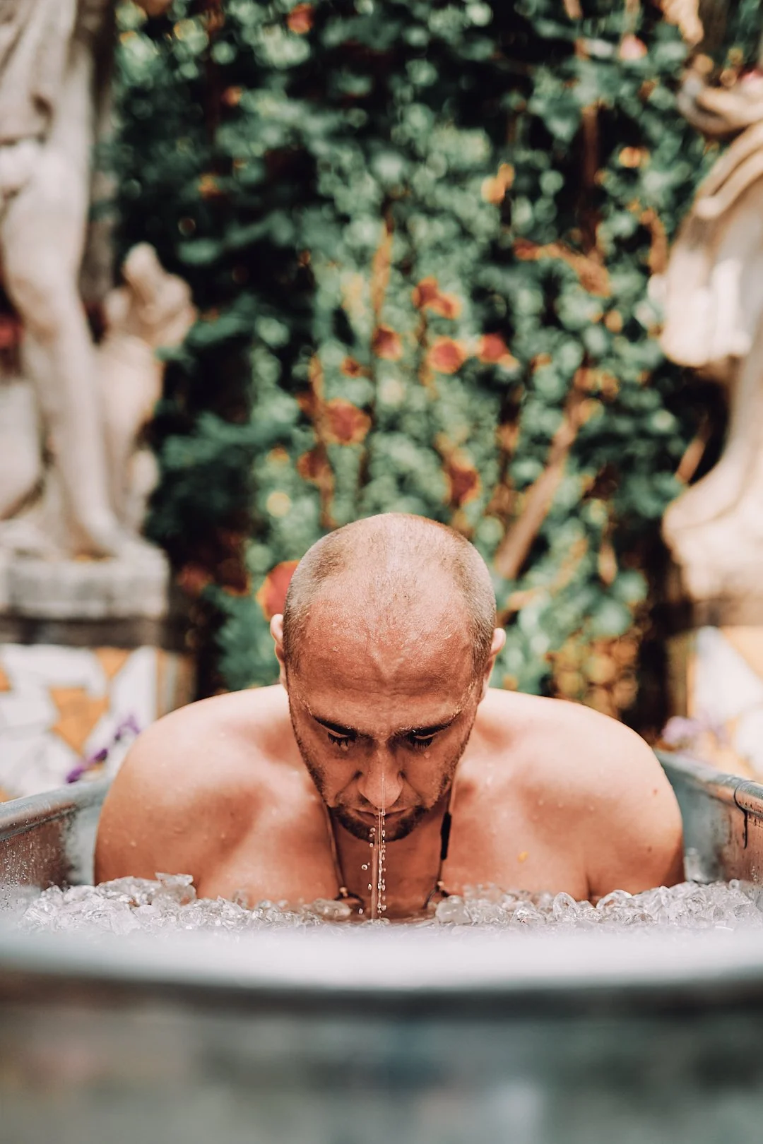 Ein Mann mit einem kahlen Kopf taucht in eine Badewanne mit Wasser und Eis, vor einem grünen, blühenden Hintergrund.