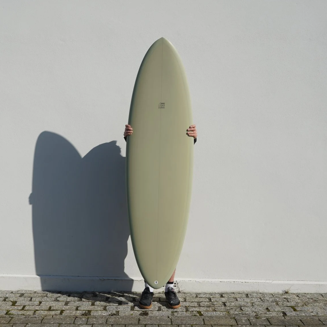 Person standing on a cobblestone ground holding an off-white surfboard vertically against a plain white wall, with a shadow cast on the wall.