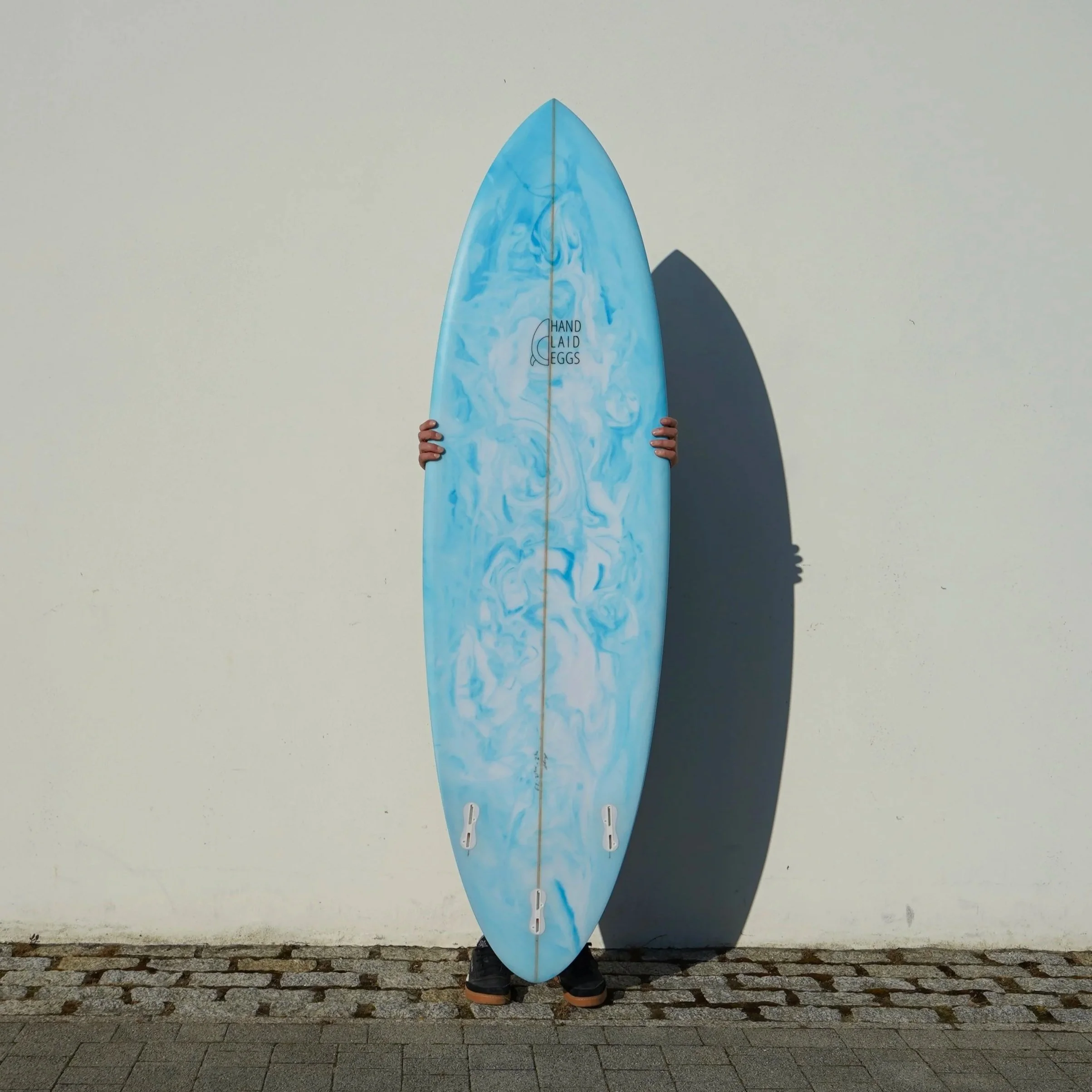 Person holding a blue and white marbled surfboard with the words 'Hand Laid Eggs' on it, standing against a light gray wall, with their shoes visible at the bottom of the surfboard.