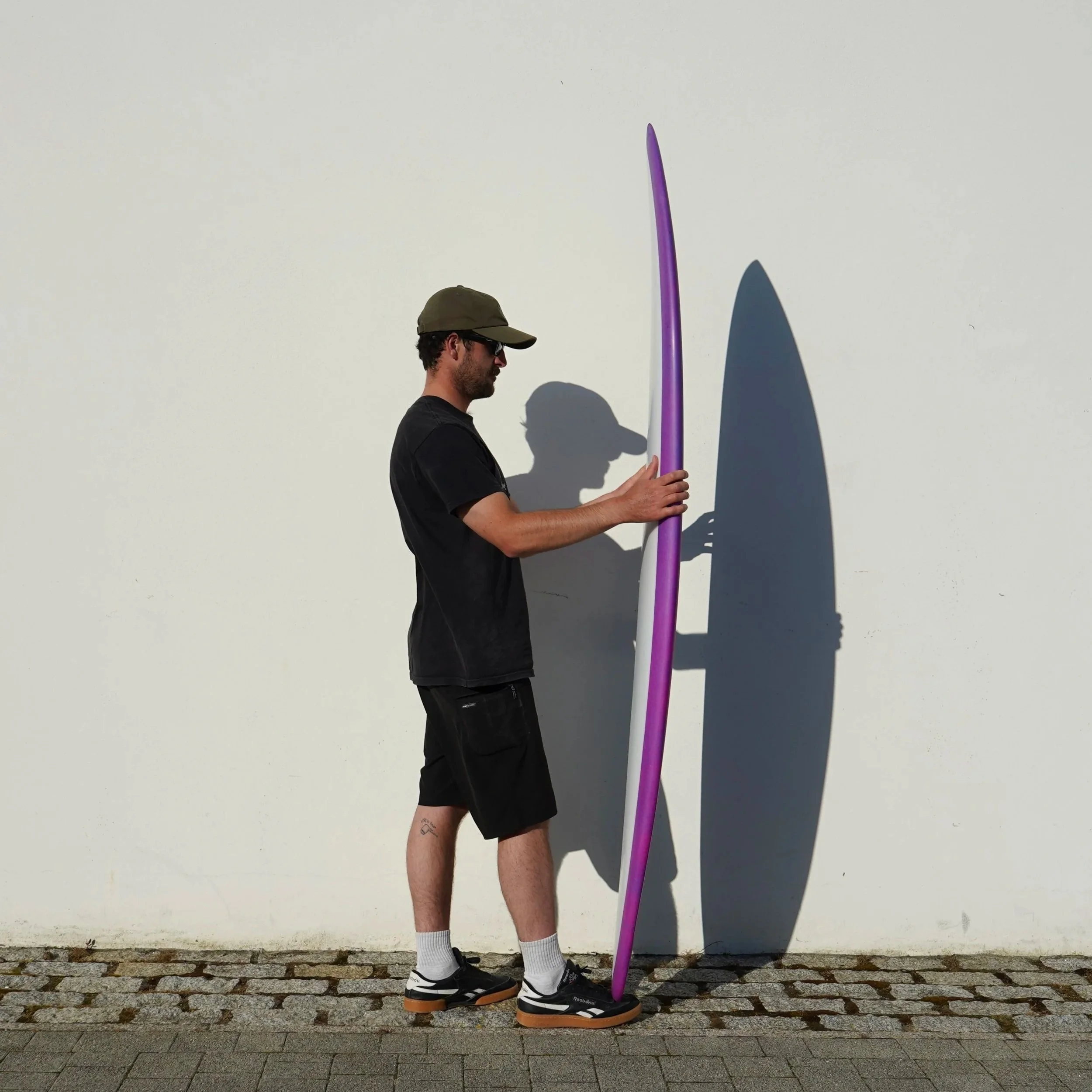 A man with dark hair, beard, and glasses wearing a green cap, black T-shirt, black shorts, white socks, and sneakers, is standing on a cobblestone sidewalk against a plain white wall. He is holding a large purple object that looks like a surfboard or