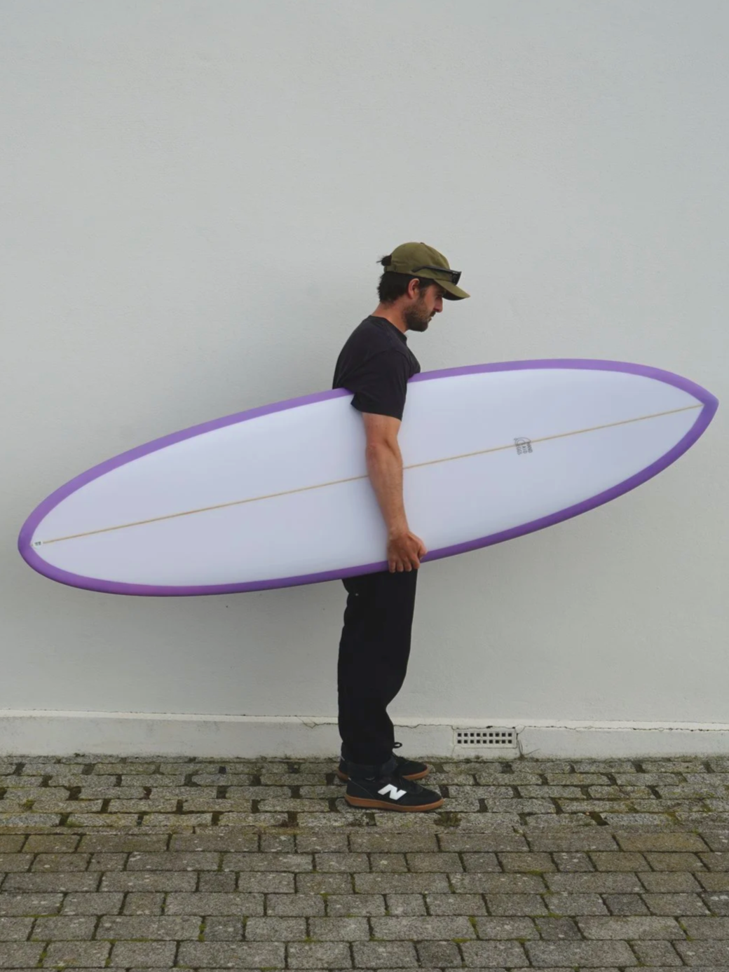 Man standing against a plain wall, holding a white surfboard with purple trim, wearing a black t-shirt, black pants, black shoes with white logos, and a green cap with sunglasses.