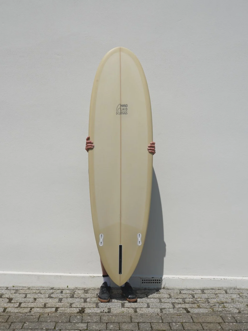 Person holding a longboard skateboard vertically against a plain white wall, feet visible at the bottom, shadow cast on the wall.