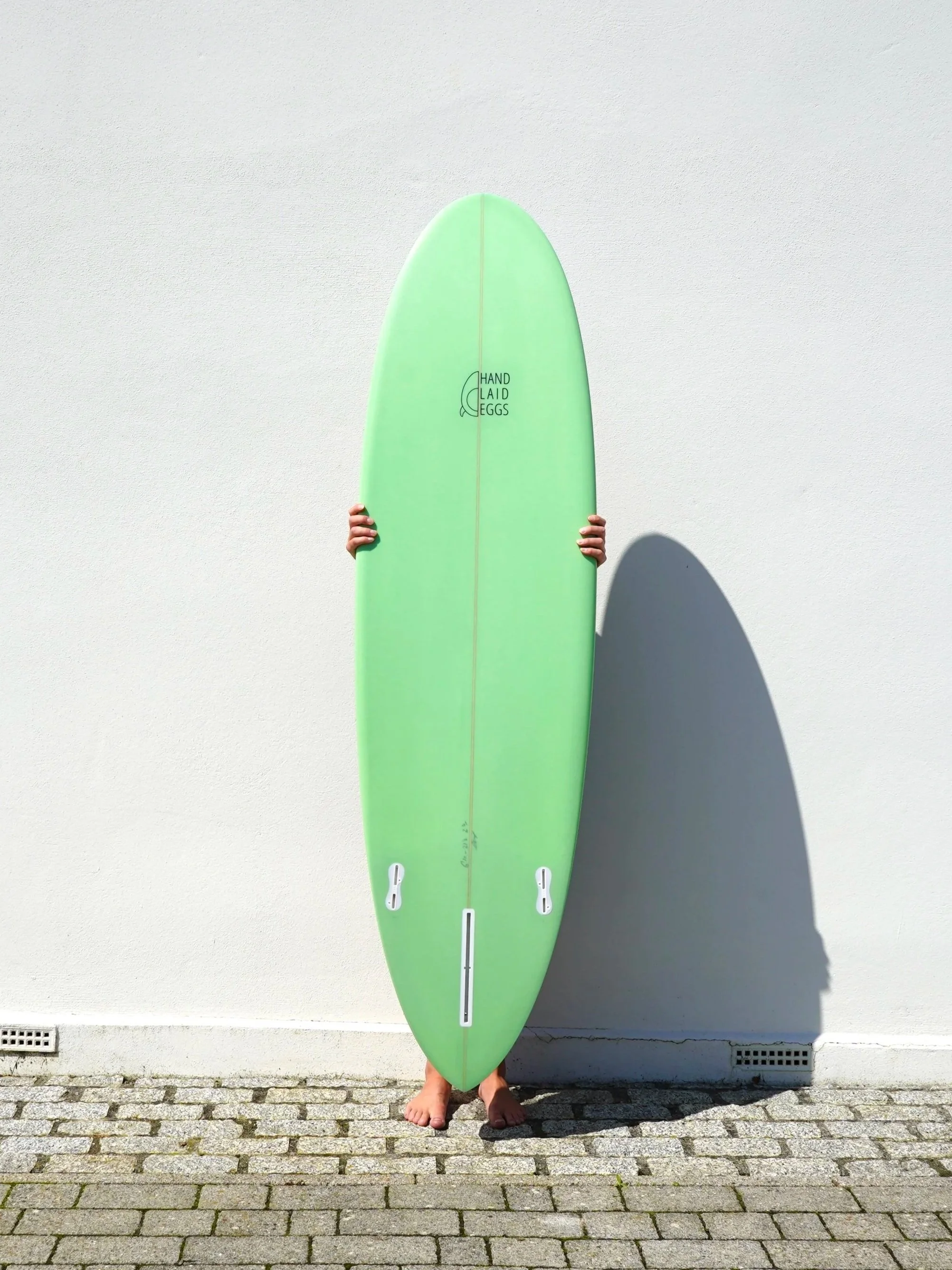 Person holding a light green surfboard upright against a plain white wall, with their feet visible on cobblestone pavement, casting a shadow on the wall.