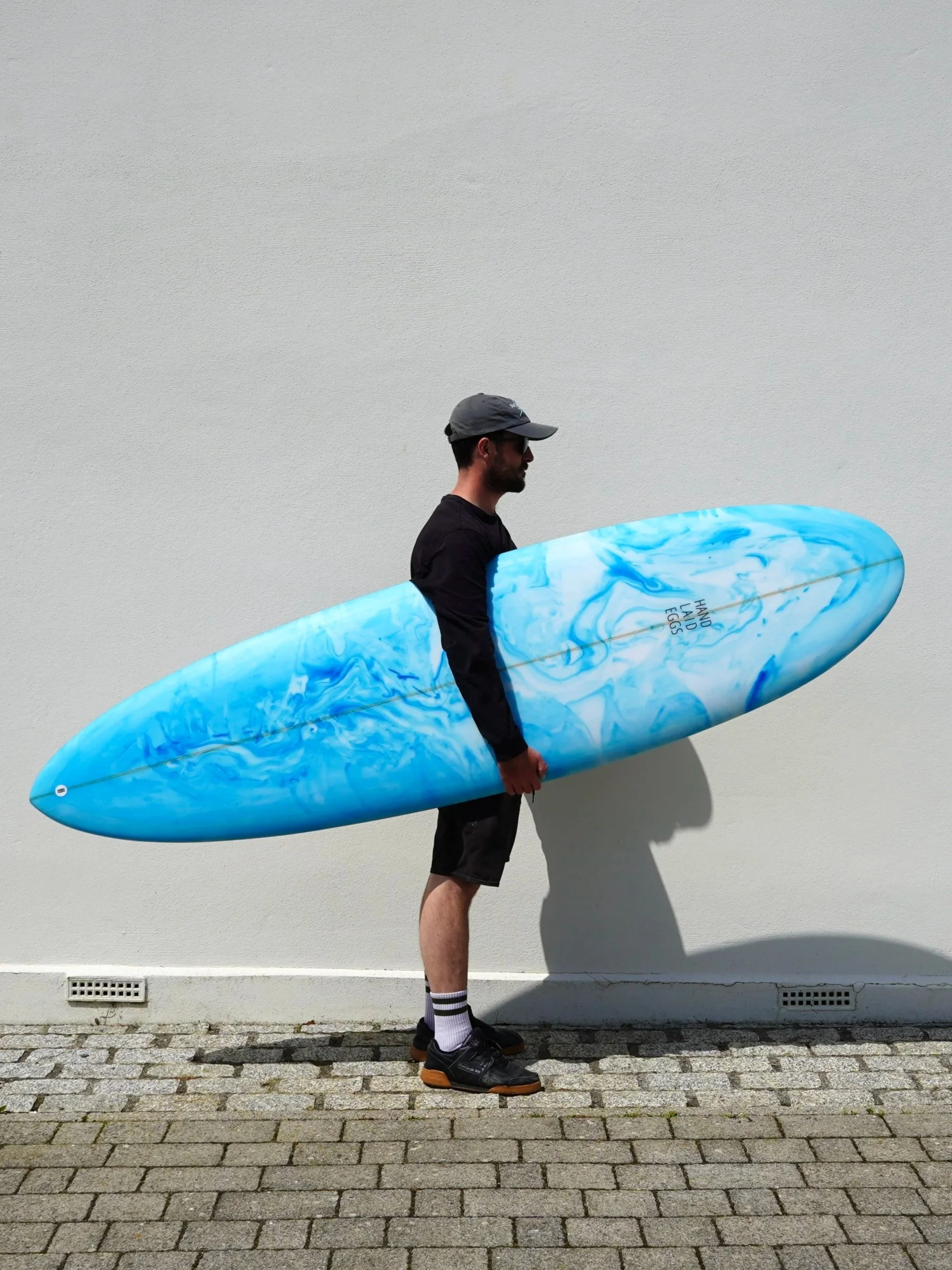 A man wearing a gray baseball cap, black sunglasses, a black T-shirt, shorts, and black sneakers holding a blue and white marbled surfboard with the words "Hand Laid Eggs" written on it, standing against a plain white wall on a cobblestone sidewalk.