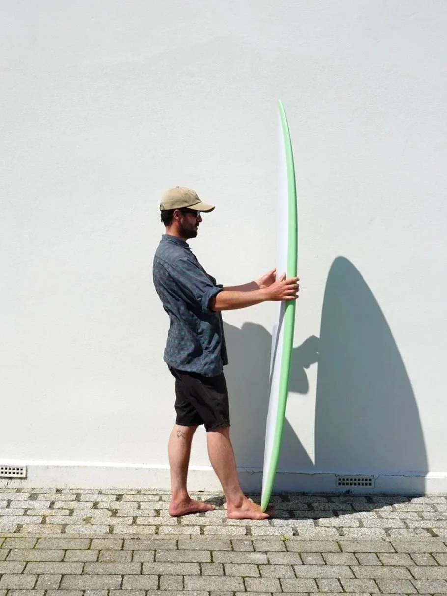 A man in a baseball cap, sunglasses, a gray shirt, and black shorts holding a surfboard with a green edge against a white wall, casting a shadow.