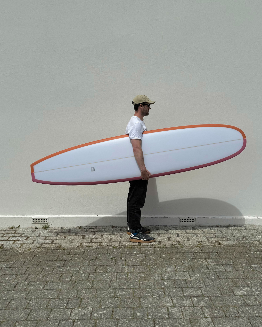 A man standing against a plain wall, holding a longboard surfboard, dressed casually in a white t-shirt, black pants, sneakers, beige cap, and sunglasses.