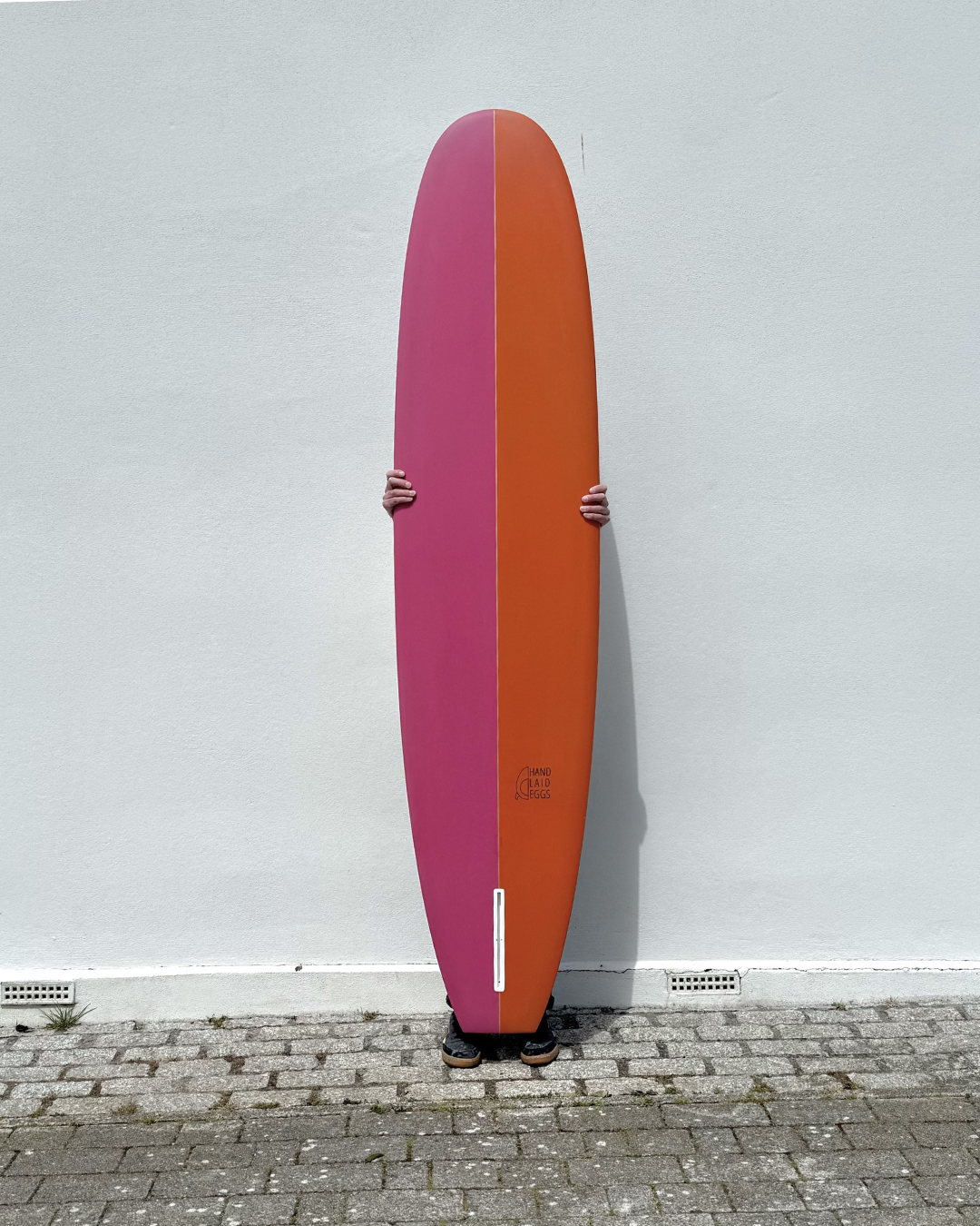 Person holding a large pink and orange longboard surfboard against a white wall.