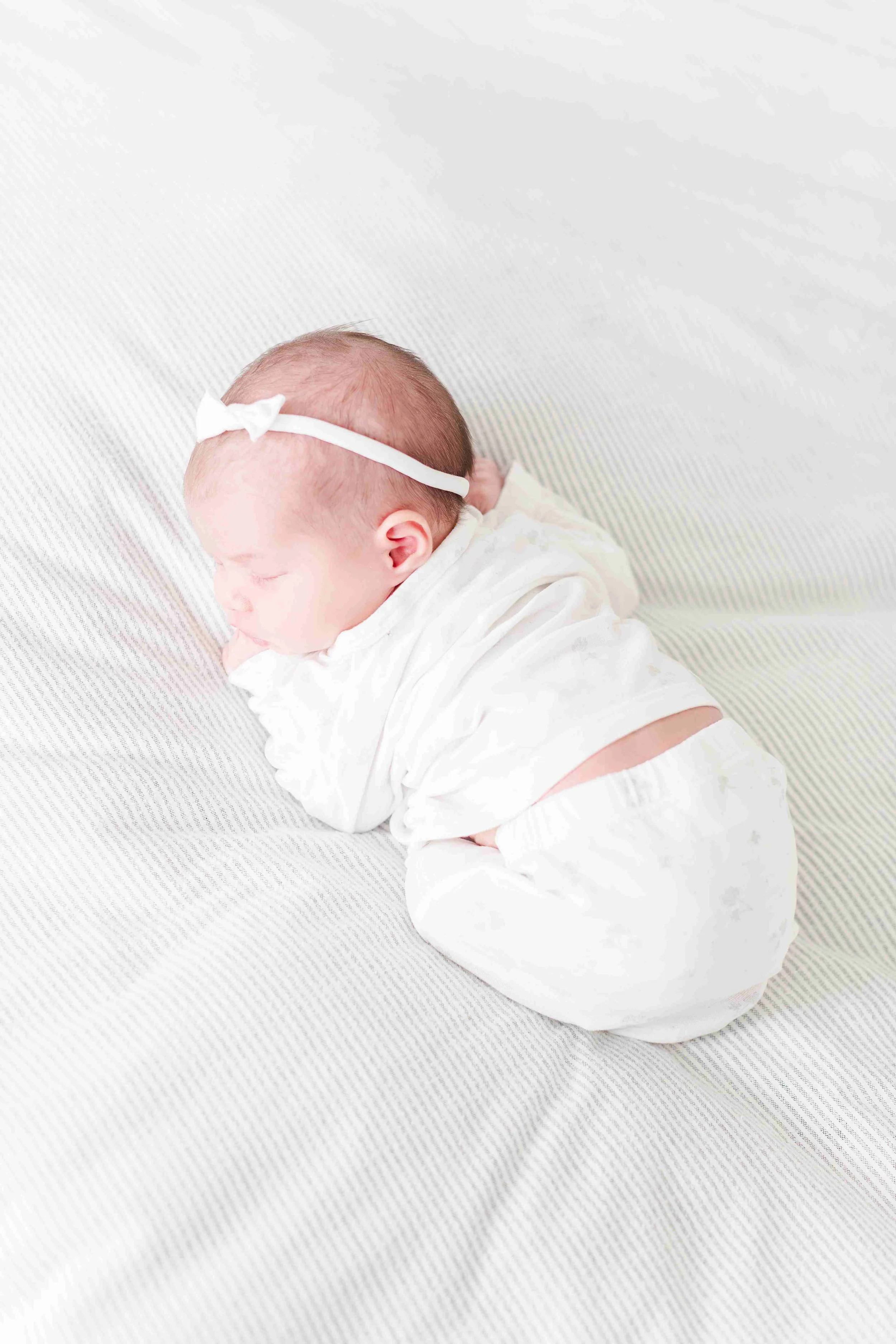 A sleeping baby dressed in white lay on a bed with a light-colored striped sheet, with a white headband with a bow on their head.