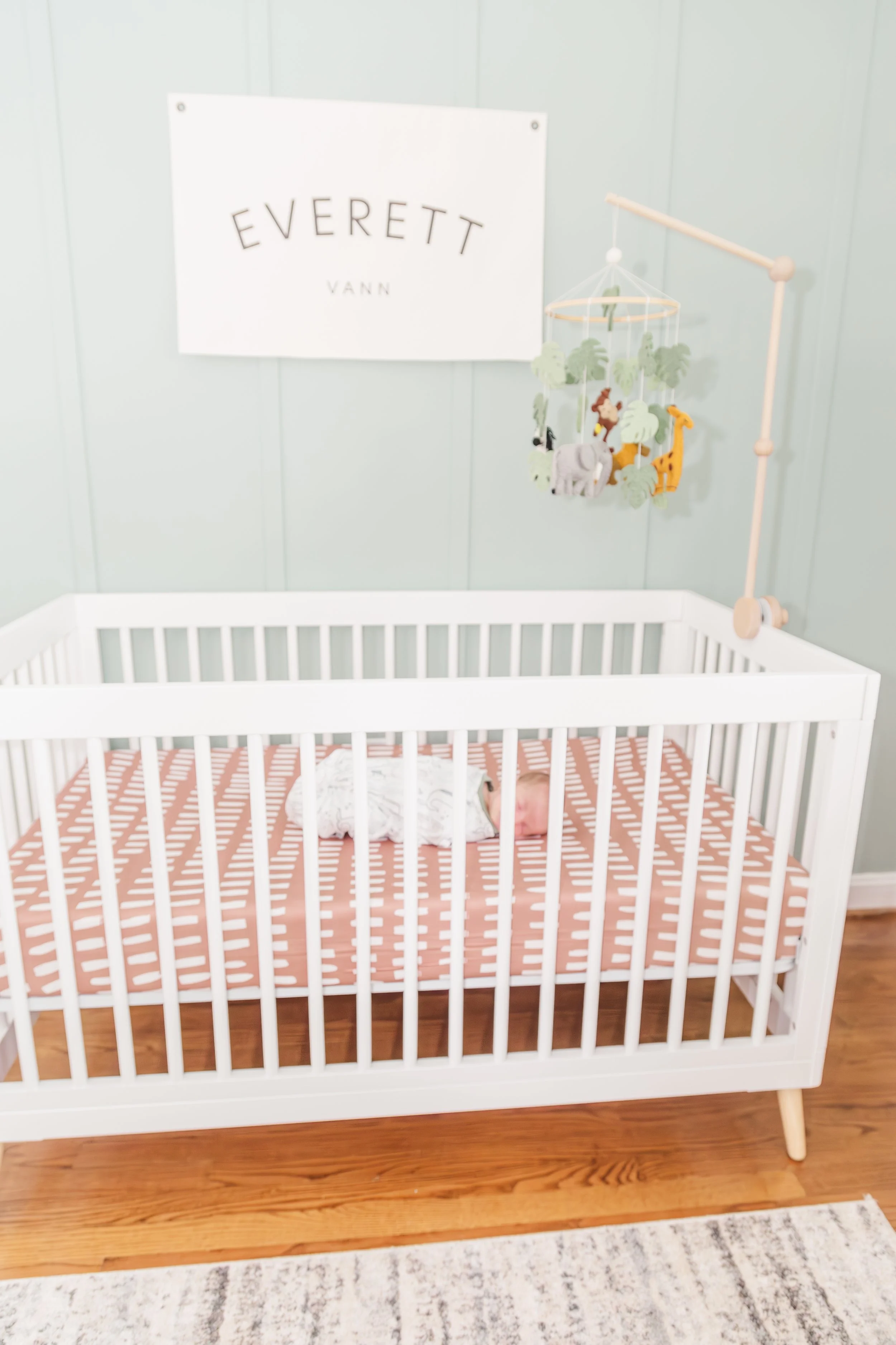A white crib with a baby sleeping inside, surrounded by a patterned pink and white sheet. Above the crib is a mobile with animal figures. A sign with text is hanging on the wall in the background.