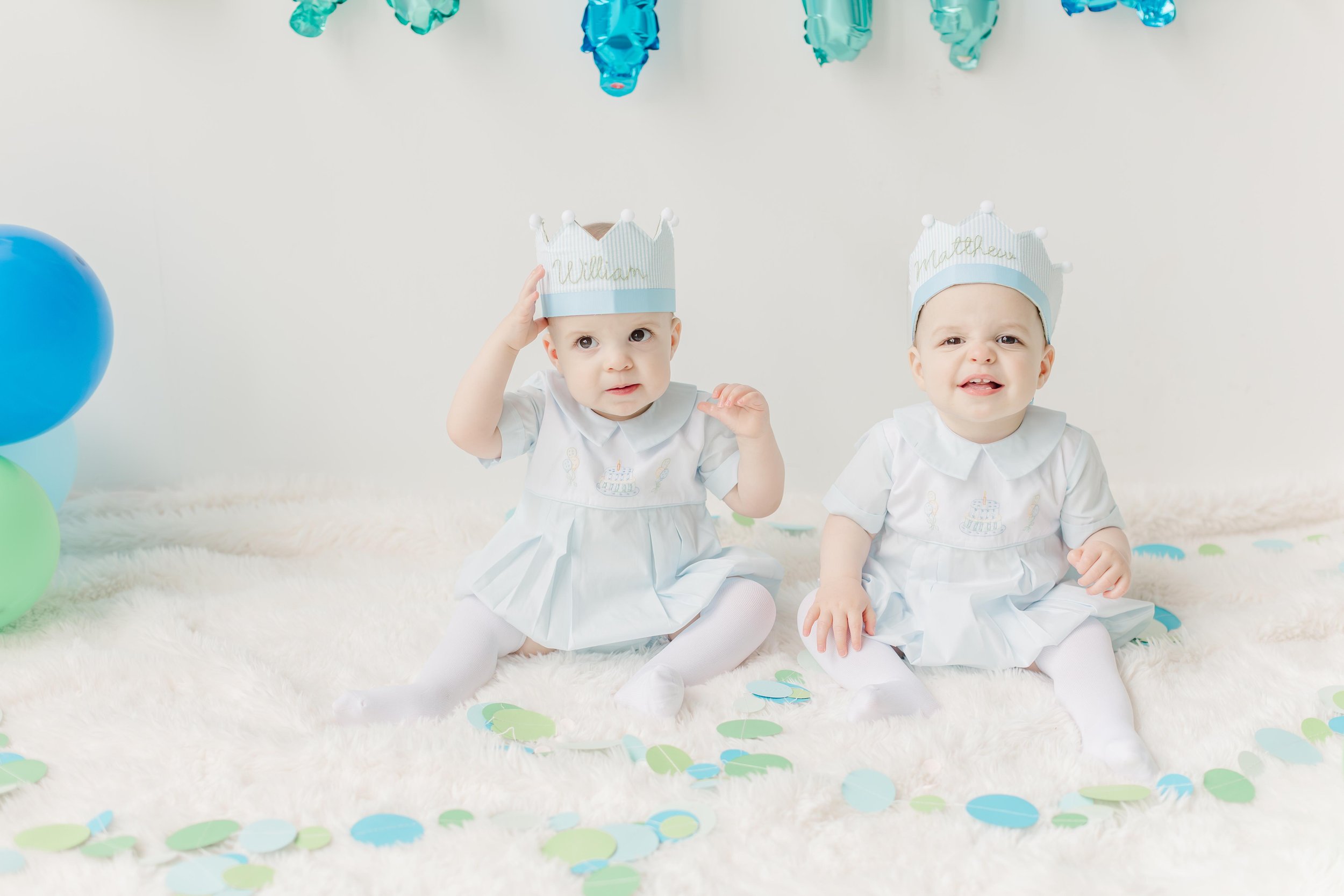 Two young children dressed in white outfits with birthday cake embroidery, wearing paper crowns with their names, sitting on a fluffy white rug surrounded by blue and green balloons and confetti, at a birthday celebration.