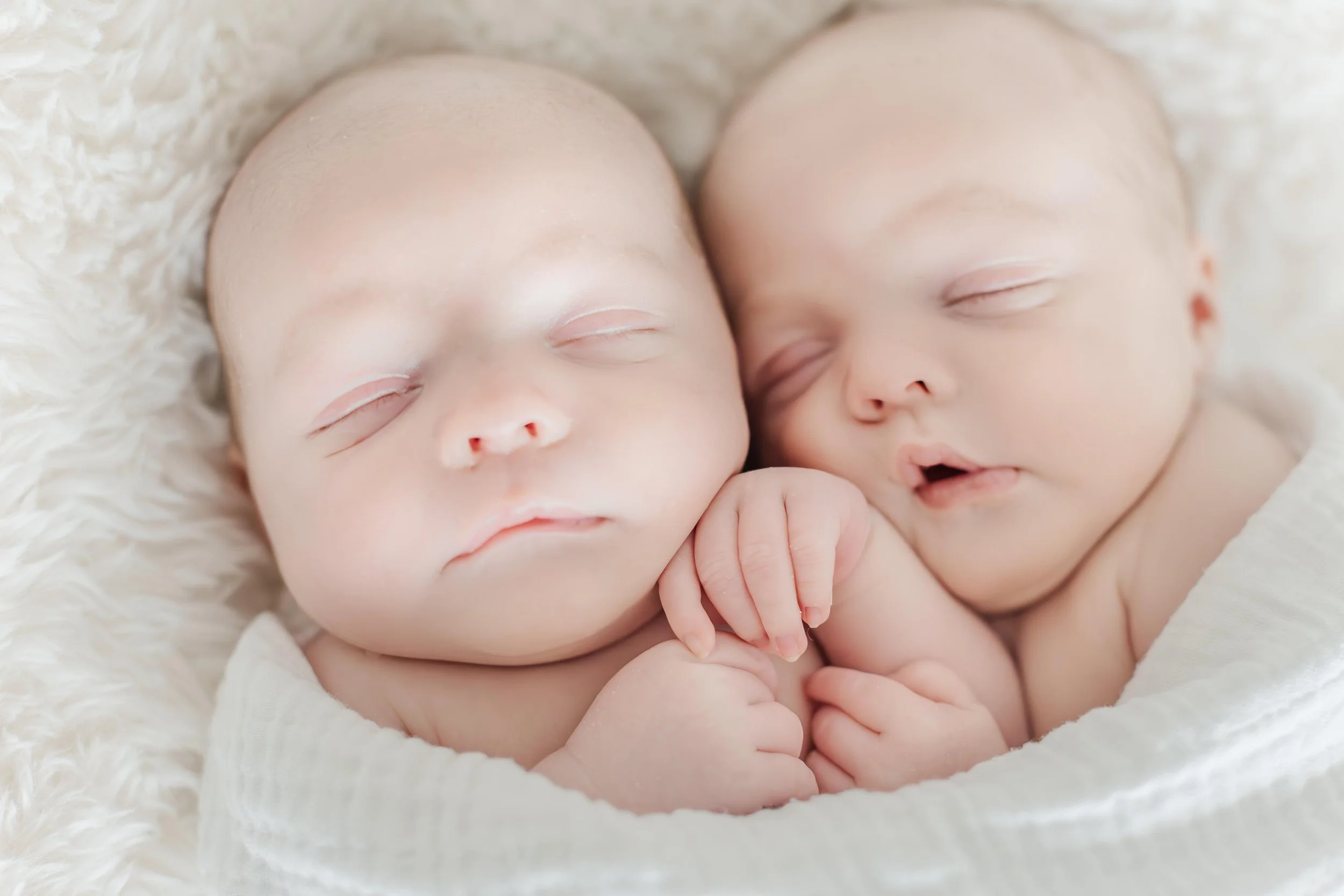 Two sleeping babies with closed eyes, resting on a fluffy white blanket, with one baby gently touching the other's hand.