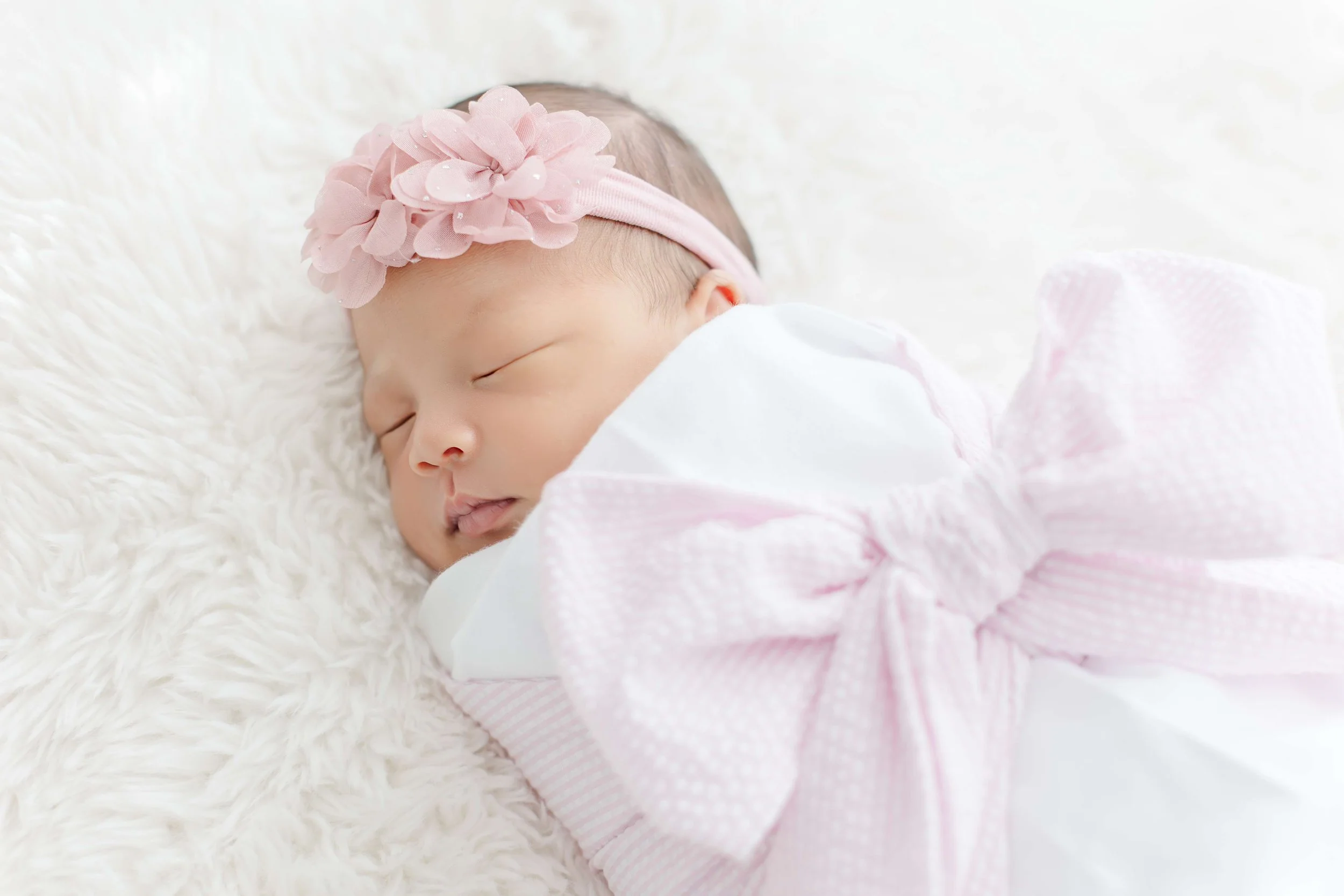 Newborn baby girl swaddled in white with a pink flower headband, eyes closed peacefully — Greenville South Carolina newborn photography