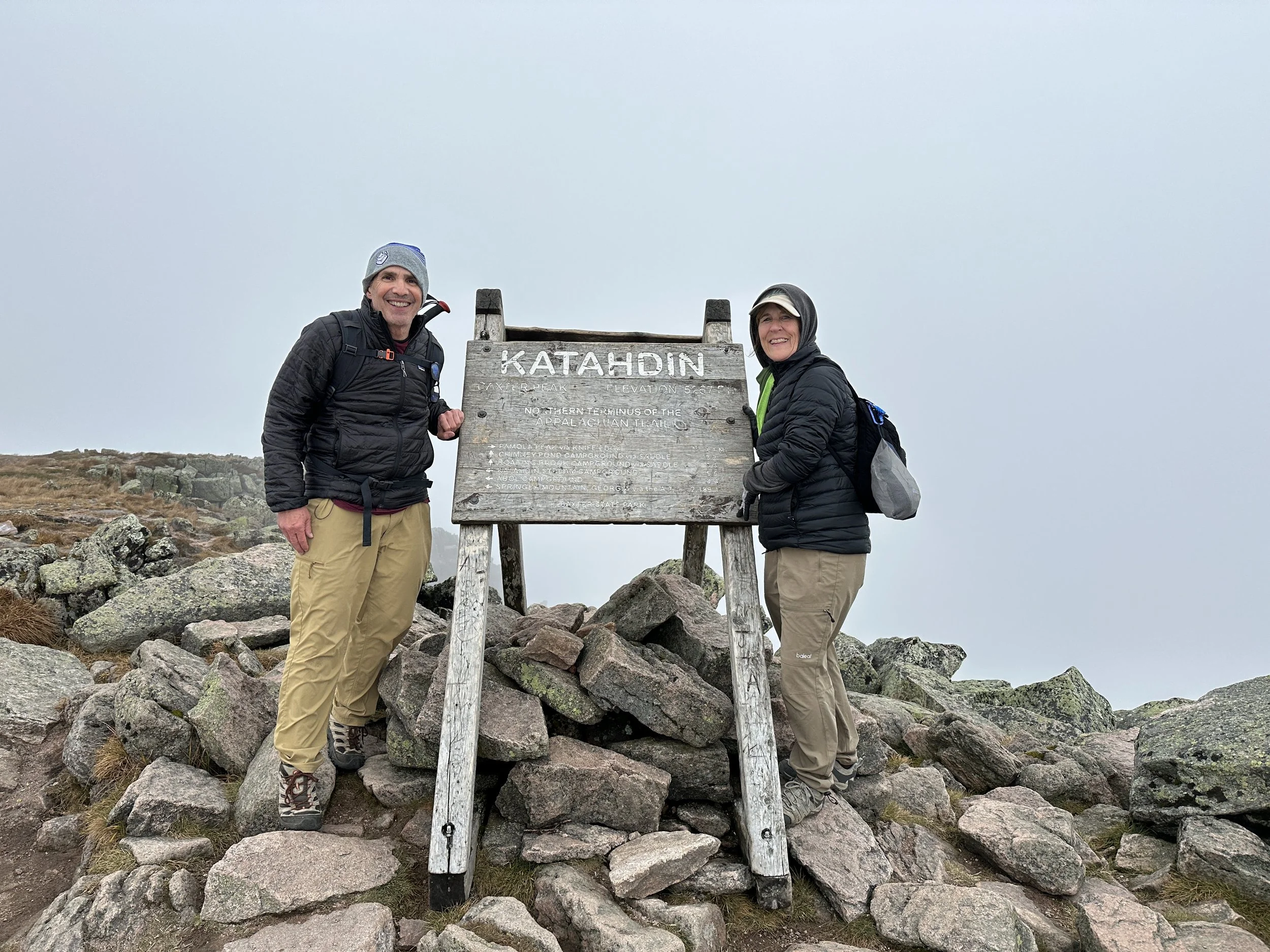 Two hikers standing next to a wooden sign that reads 'KATAHDIN' at the summit, surrounded by rocks with a foggy sky in the background.