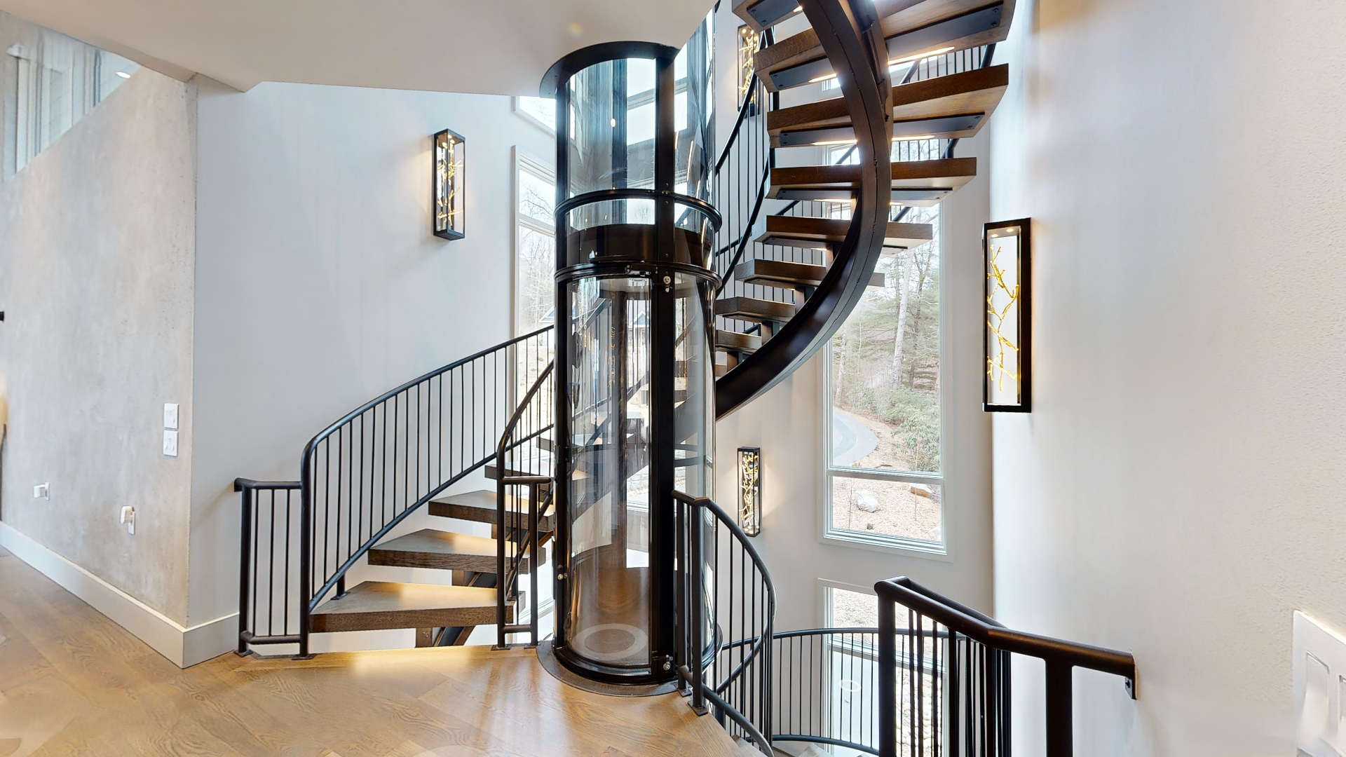 Interior view of a modern house staircase with a curved wooden staircase, glass elevator, and large windows showing trees outside.