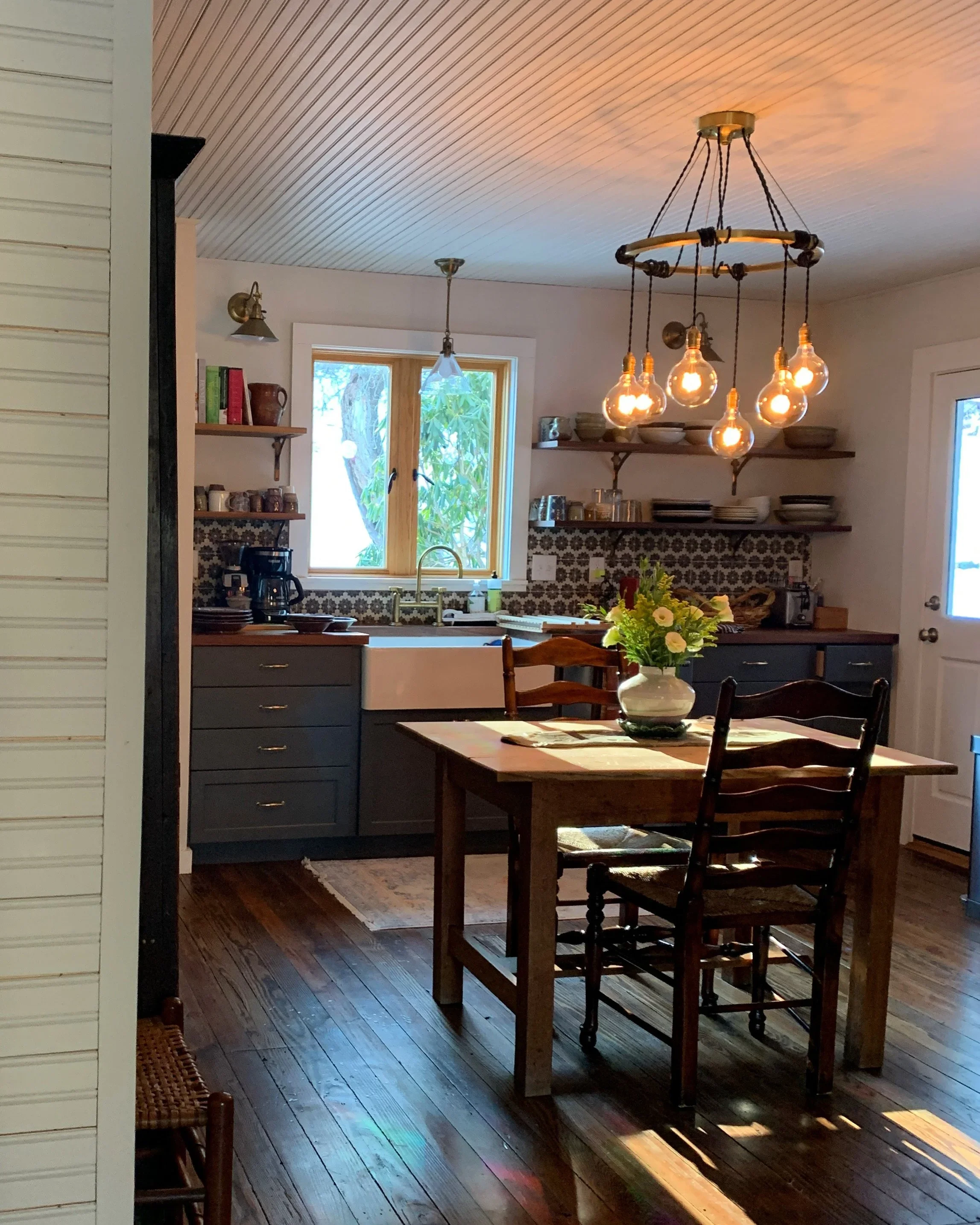 A cozy kitchen and dining area with wooden flooring, a wooden dining table with a flower vase, and a rustic chandelier with exposed bulbs. Kitchen cabinets are dark gray with open shelves containing dishes and kitchenware, and a window above the sink reveals greenery outside.