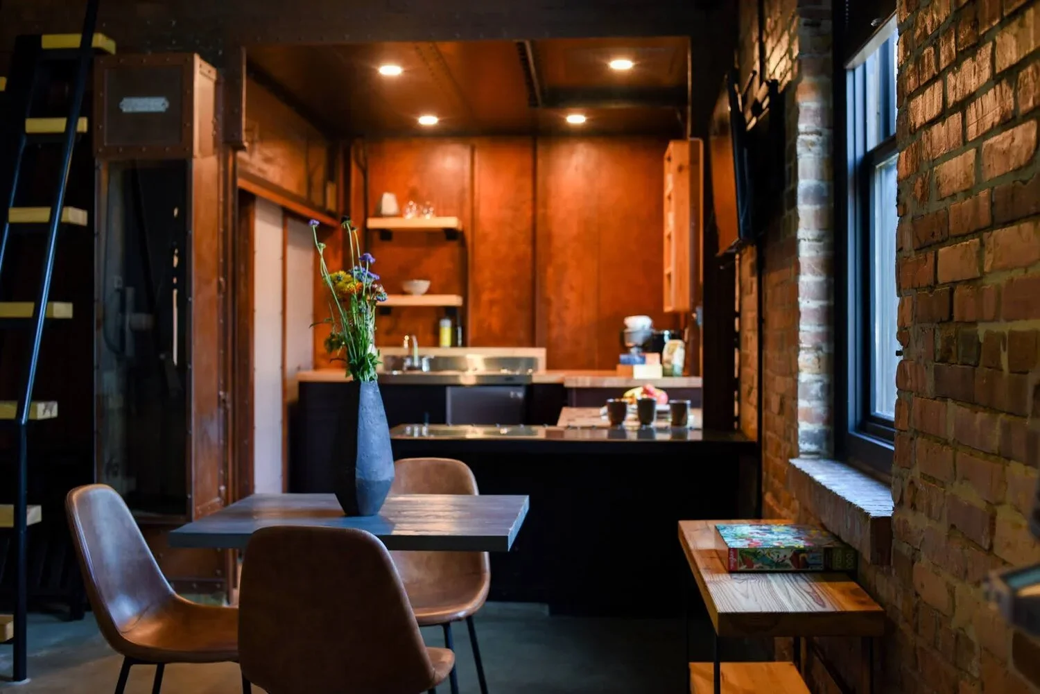 Interior of a cozy, rustic-style kitchen and dining area with wood paneling, brick walls, a black table with brown chairs, a vase with flowers, and a window letting in natural light.