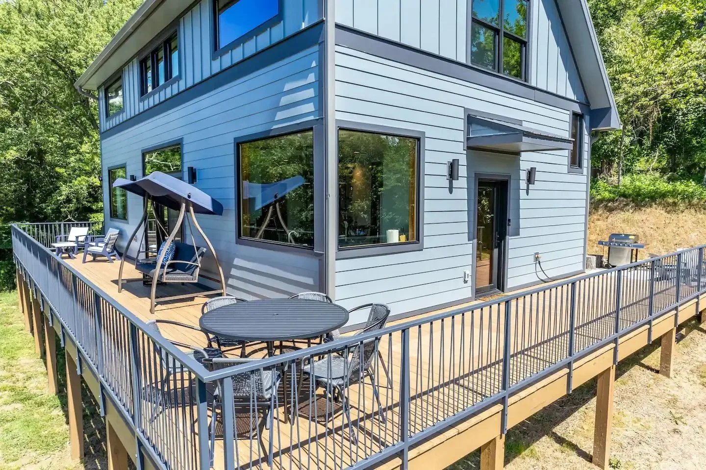 A modern two-story house with a large wooden deck, outdoor seating, and shaded swing, surrounded by green trees.